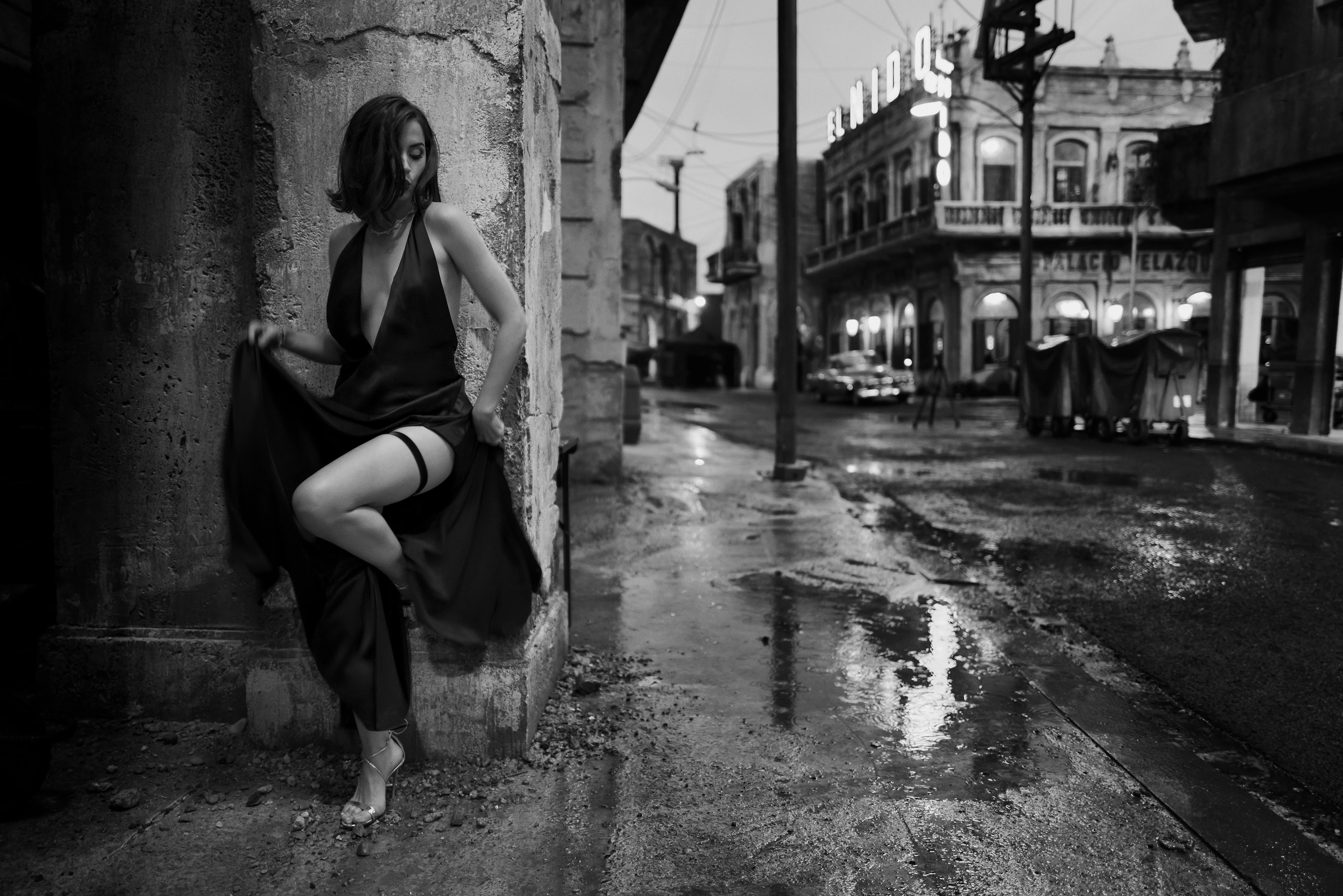 Black-and-white shot: A slender woman wearing a low-cut evening gown leans against a crumbling corner of a building at night, lifting her knee as if she were dancing and exposing her leg as puddles of rain reflect the empty street scene.