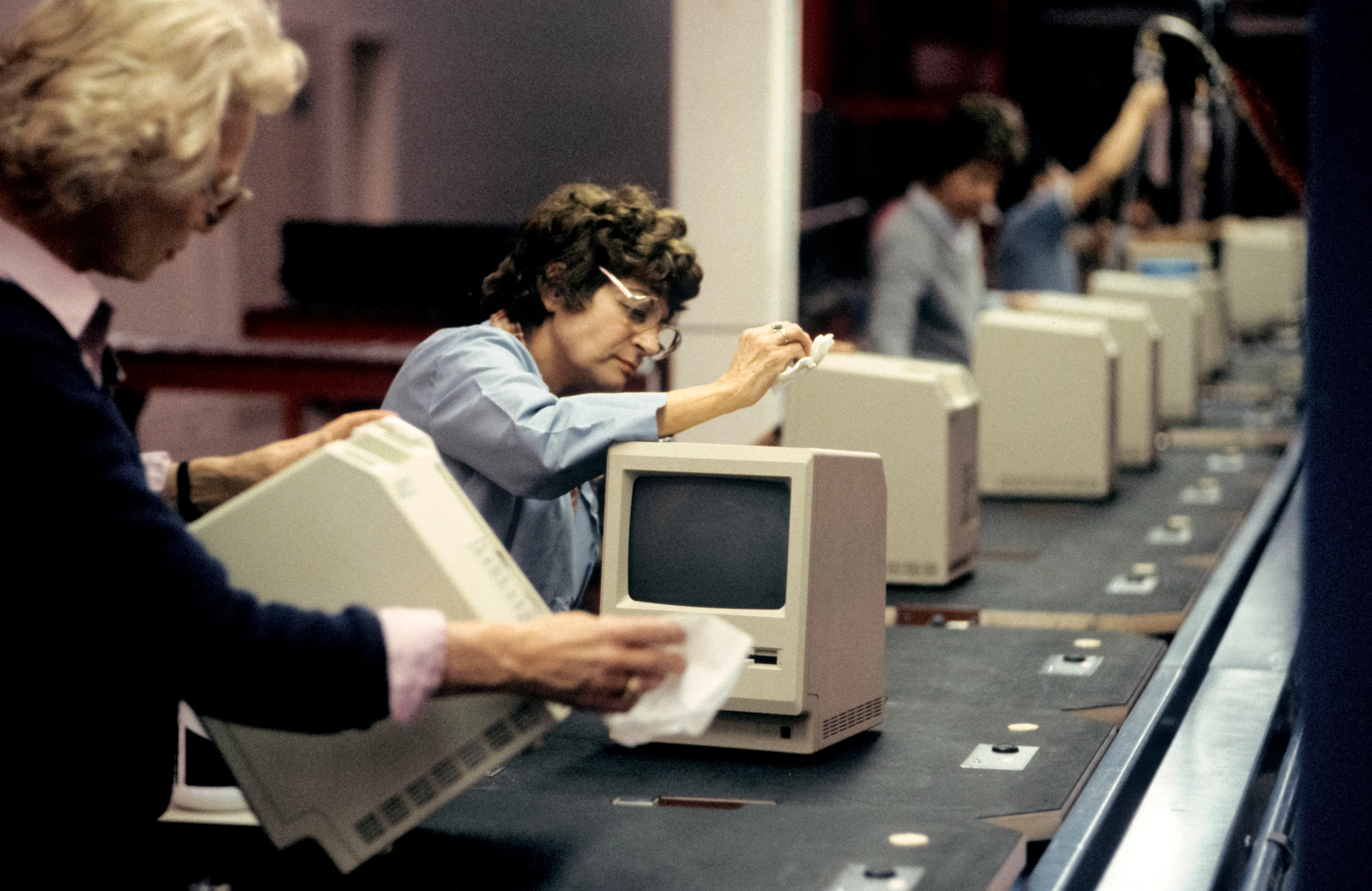 Diverse donne davanti a un nastro trasportatore controllano i computer Macintosh e li puliscono con un panno.
