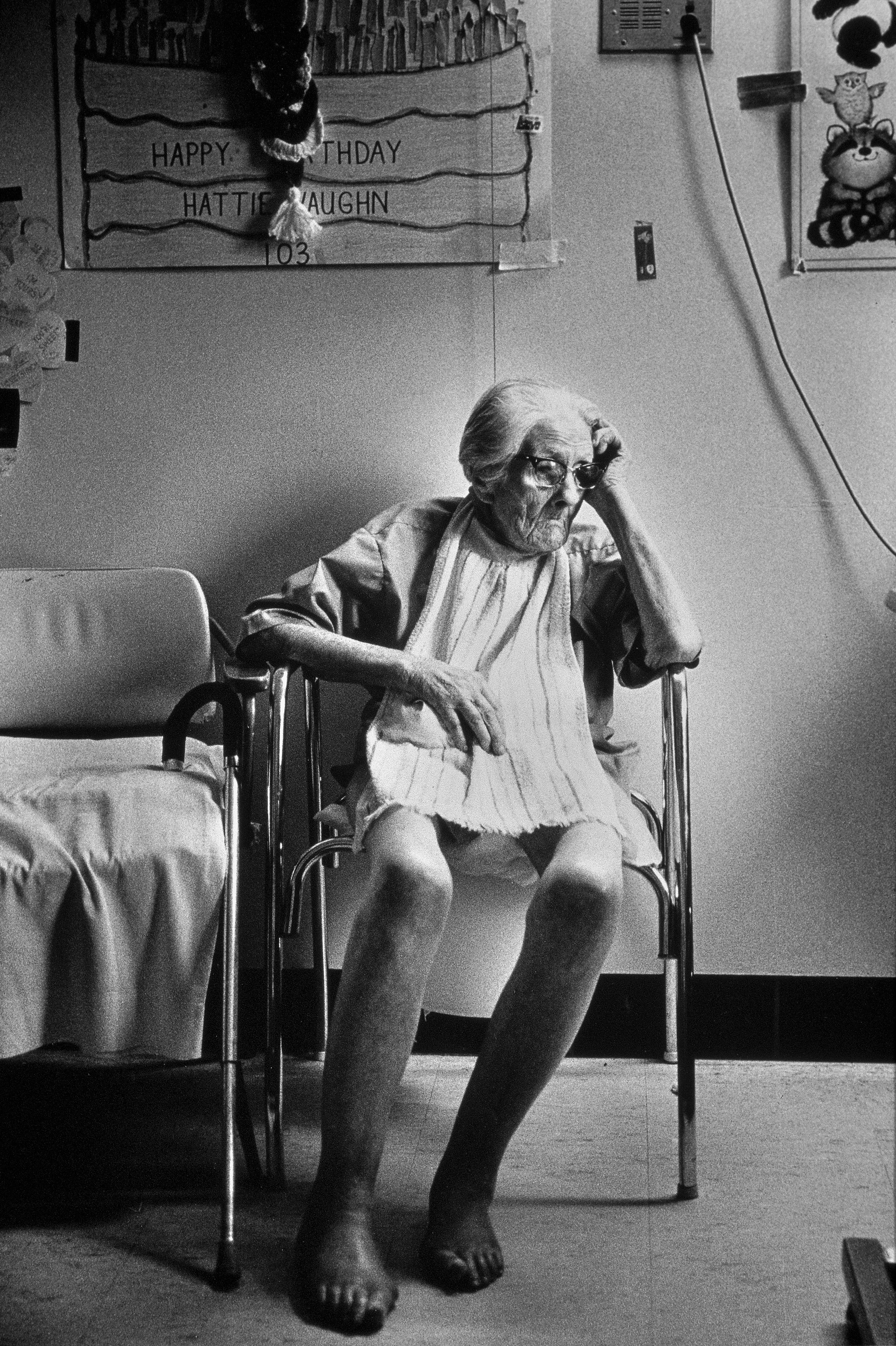 Ritratto in bianco e nero di un’anziana signora in una casa di riposo, seduta pensierosa su una sedia.