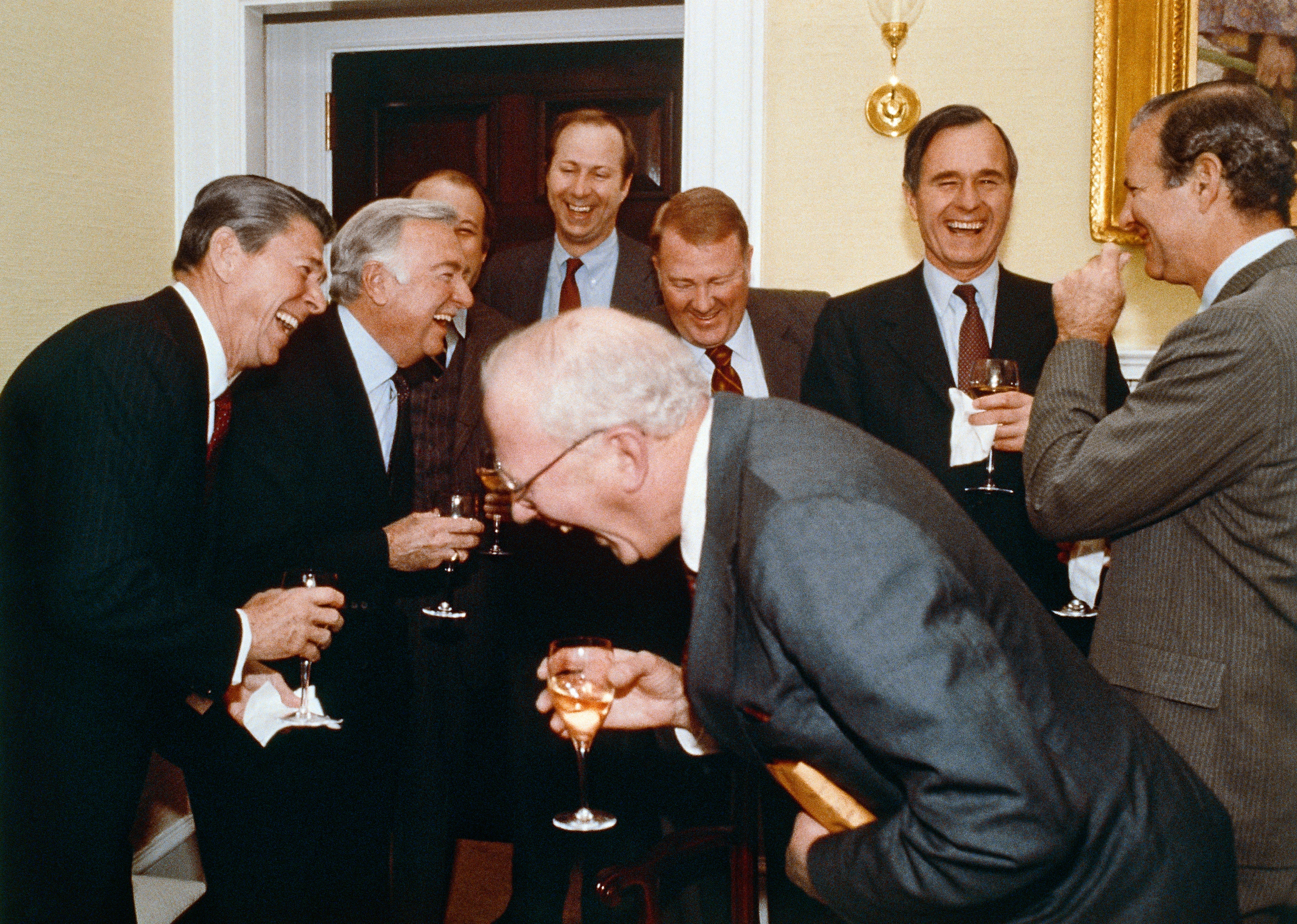 Ronald Reagan, George Bush e altri uomini ridono di cuore tenendo in mano bicchieri di spumante.