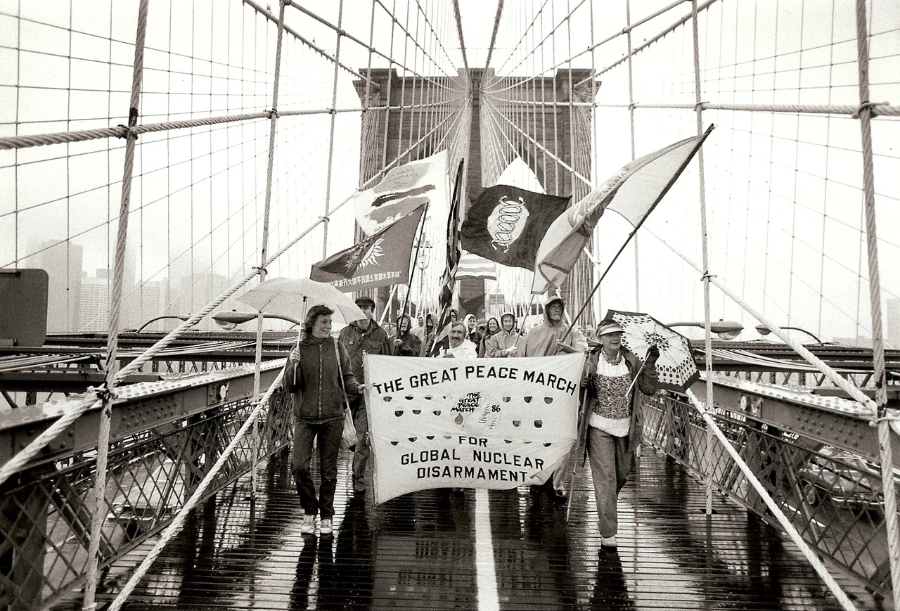 Foto in bianco e nero del fronte di un gruppo di manifestanti che attraversano il ponte di una grande città con bandiere e striscioni.