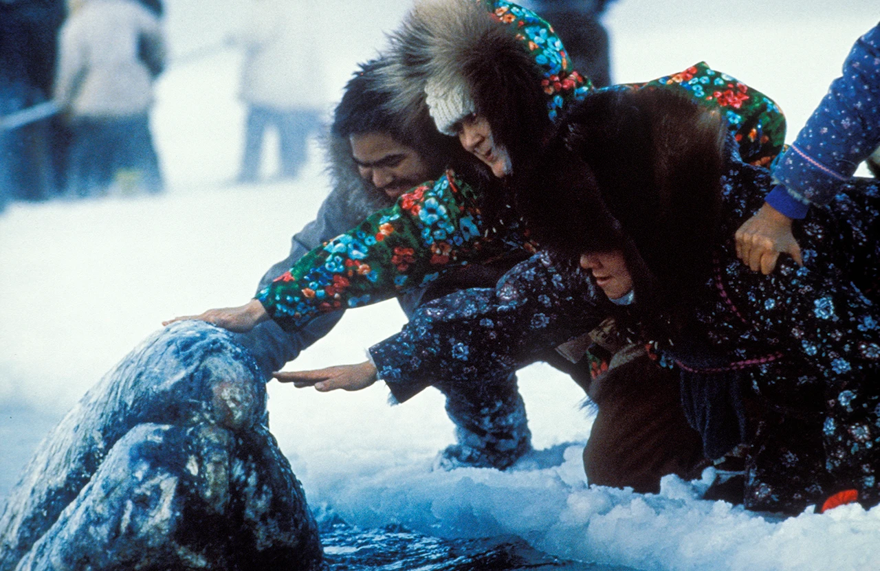 Primo piano di un gruppo di indigeni nella neve che accarezza una balena bianca la cui testa spunta dall’acqua ghiacciata.