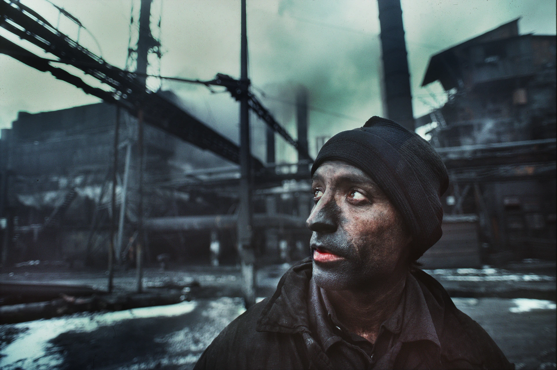 Close-up of a worker with a soot-covered face standing before a smoking industrial plant.