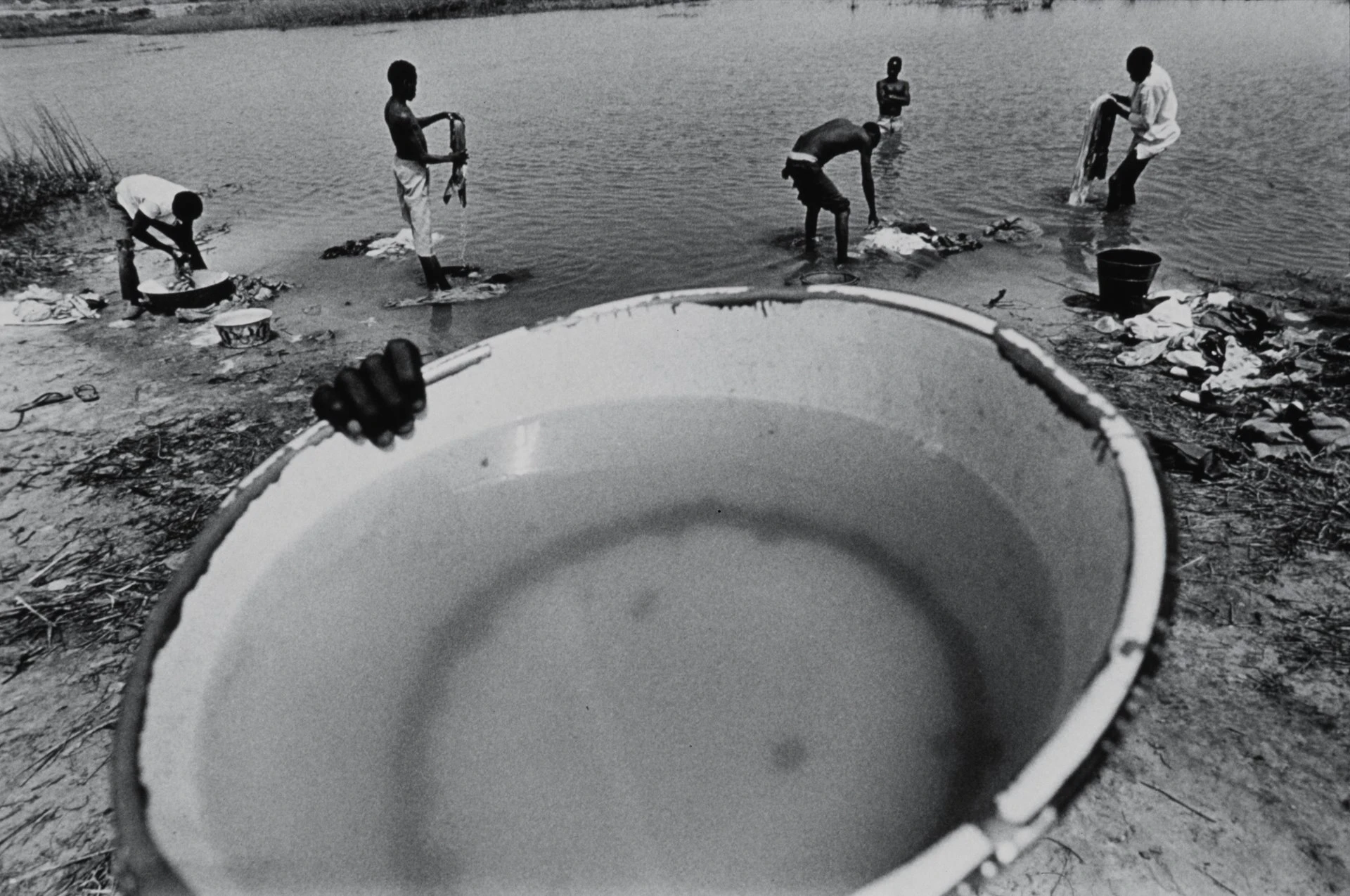 Black and white image of a waterhole in northern Nigeria, where men wash clothes in the background; in the foreground, a large washing bowl is held up towards the camera.