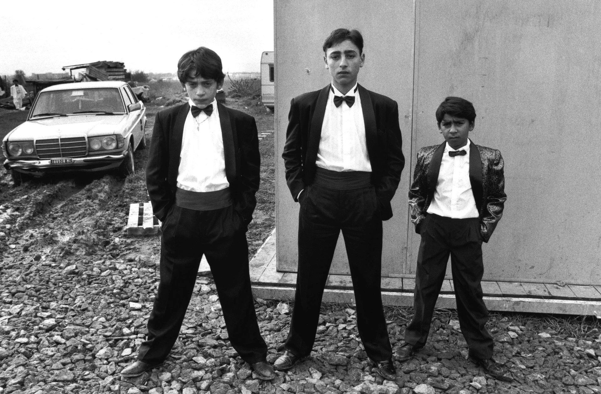 Black and white image of three Roma boys dressed smartly in suits, striking poses within a caravan estate.