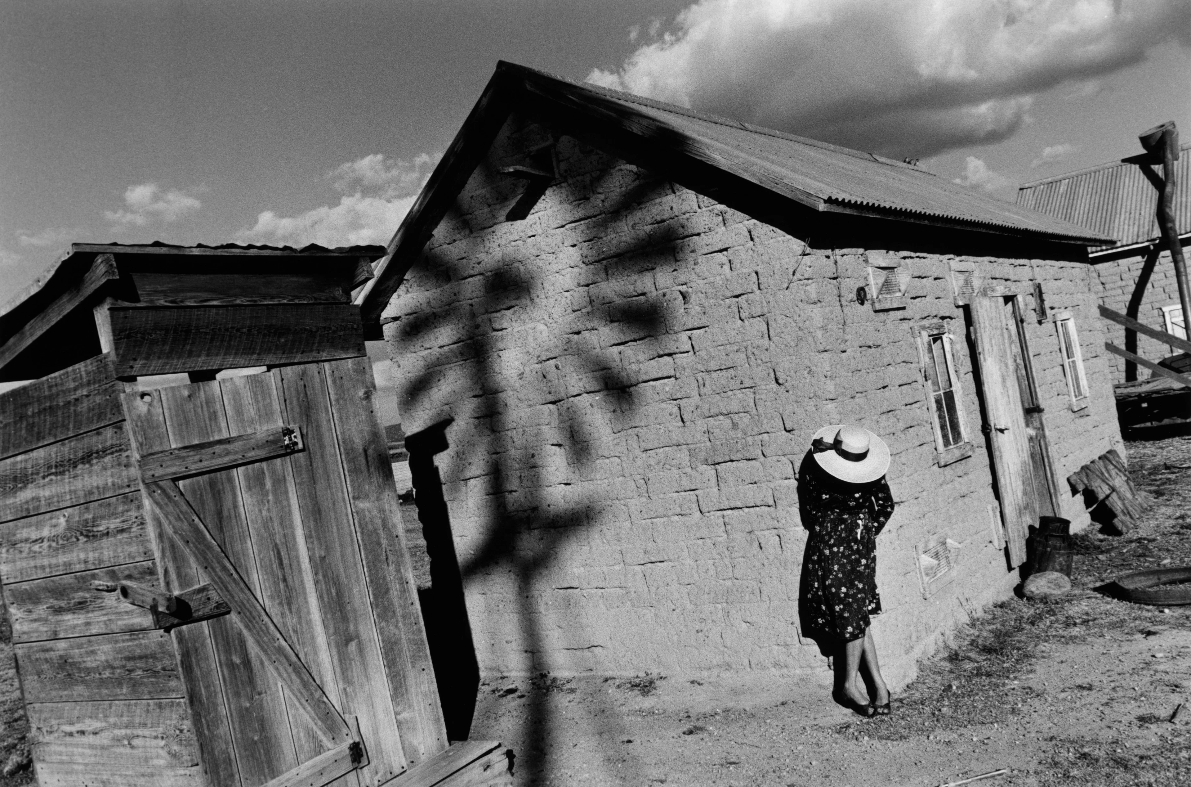 Elaborate black and white composition of a Mennonite lady in a hat posing in bright sunlight before a house. On the wall of the neighbouring building, the distinct shadow of a windmill stretches across the surface.