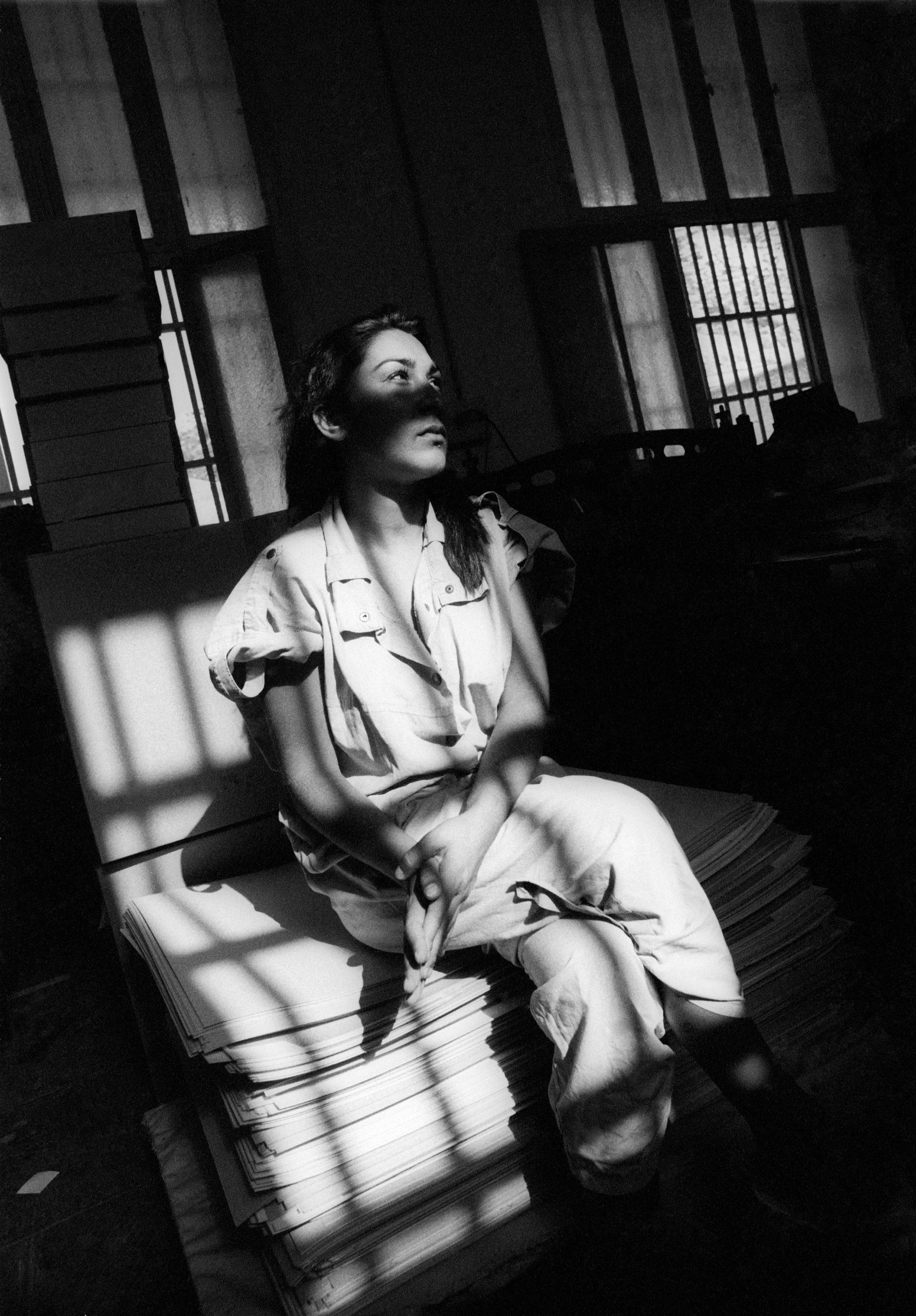 Black and white image of a woman seated with folded hands in the cone of light cast by a barred prison window, waiting.