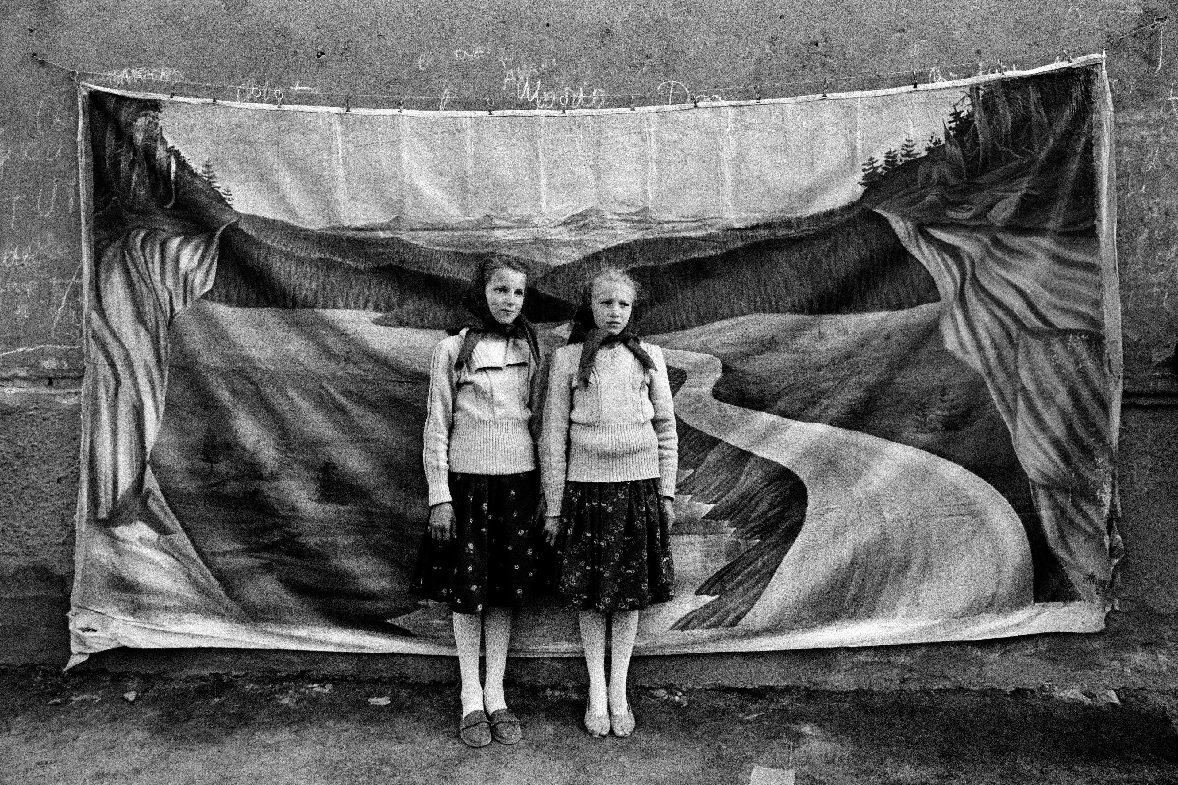 Black and white image of two girls posing in a dressy partner look before a landscape painting rendered on a sheet, which hangs against a drab, grey wall.