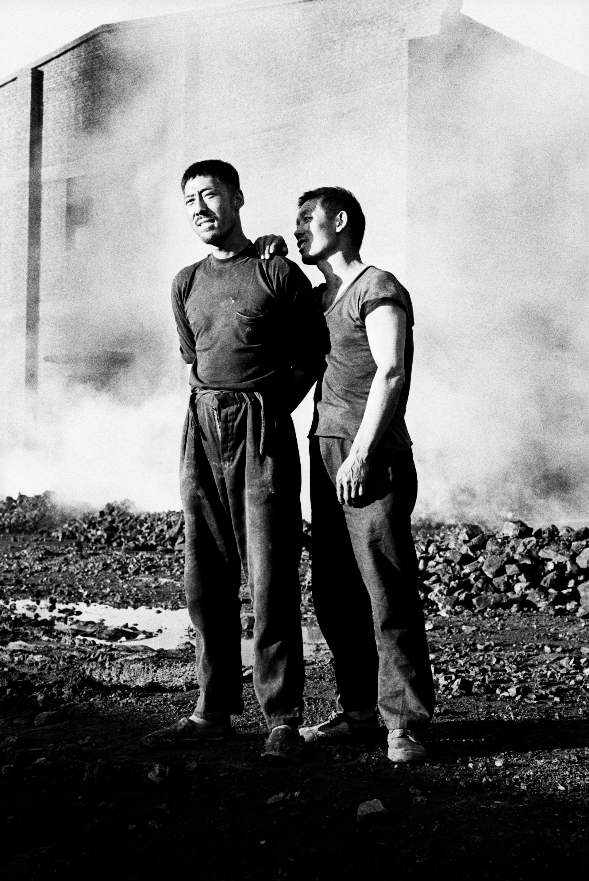 兩名面帶微笑的中國男子站在碎石街上的黑白肖像，背後是籠罩在霧氣中的工業建築。