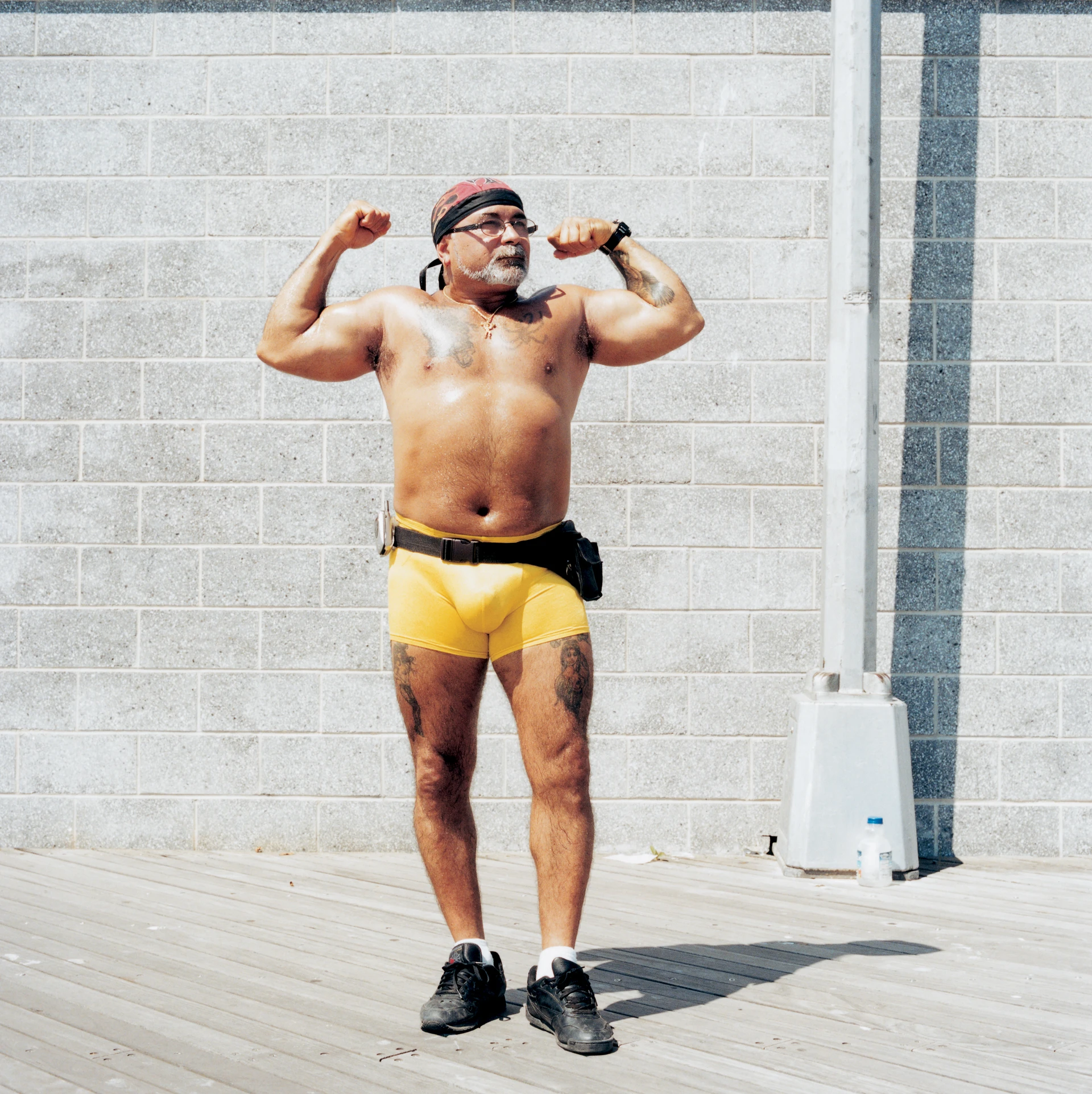 Ein oberkörperfreier, mit enger gelber Sporthose bekleideter Mann posiert für ein Porträt in der Sonne vor einer Steinmauer und zeigt seine Armmuskeln.