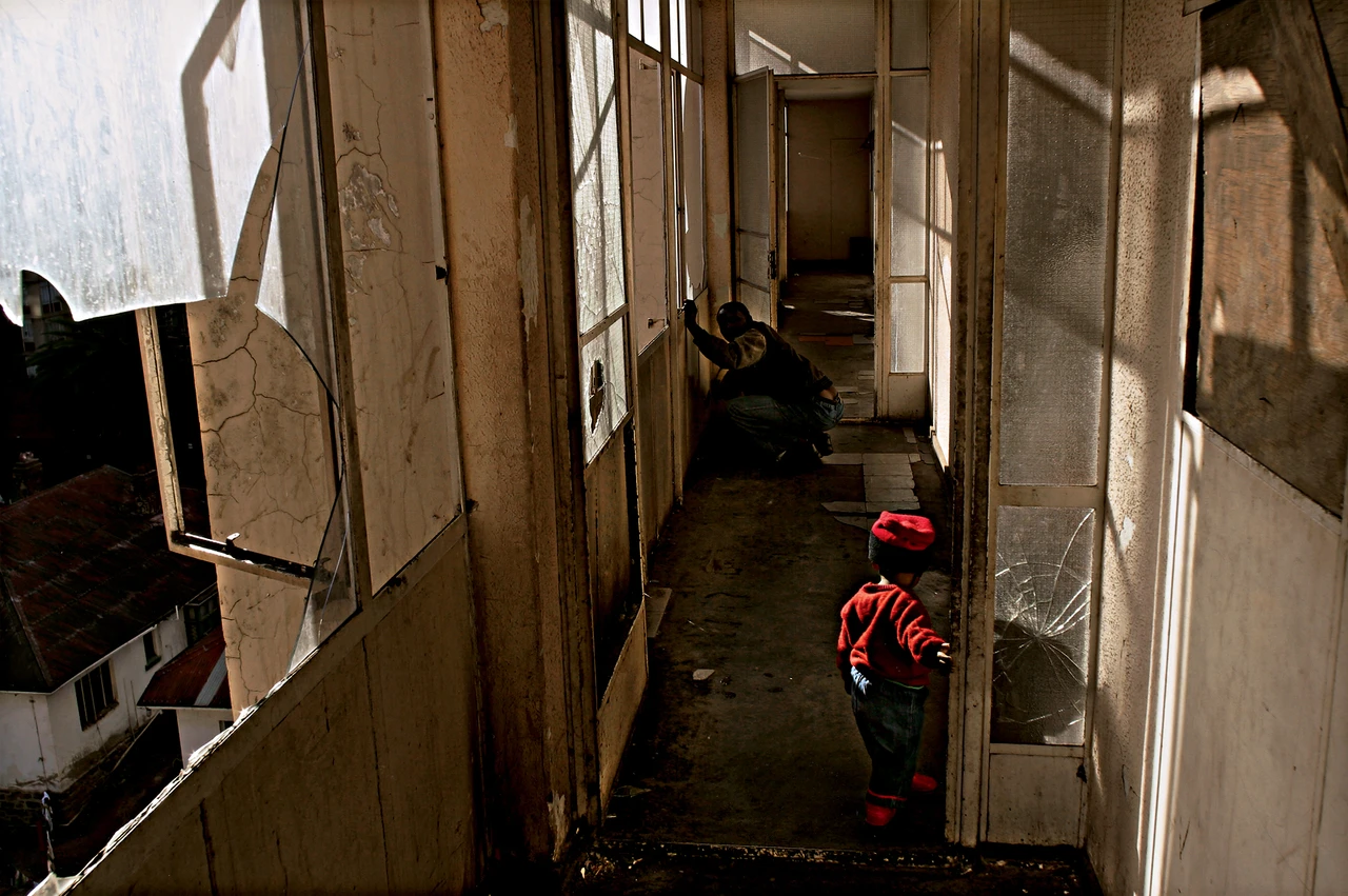 視線延伸至約翰尼斯堡一棟住宅的外廊，所有窗戶都已破碎，一個幼童沿著走廊行走，背景處一名男子蜷縮著身體。