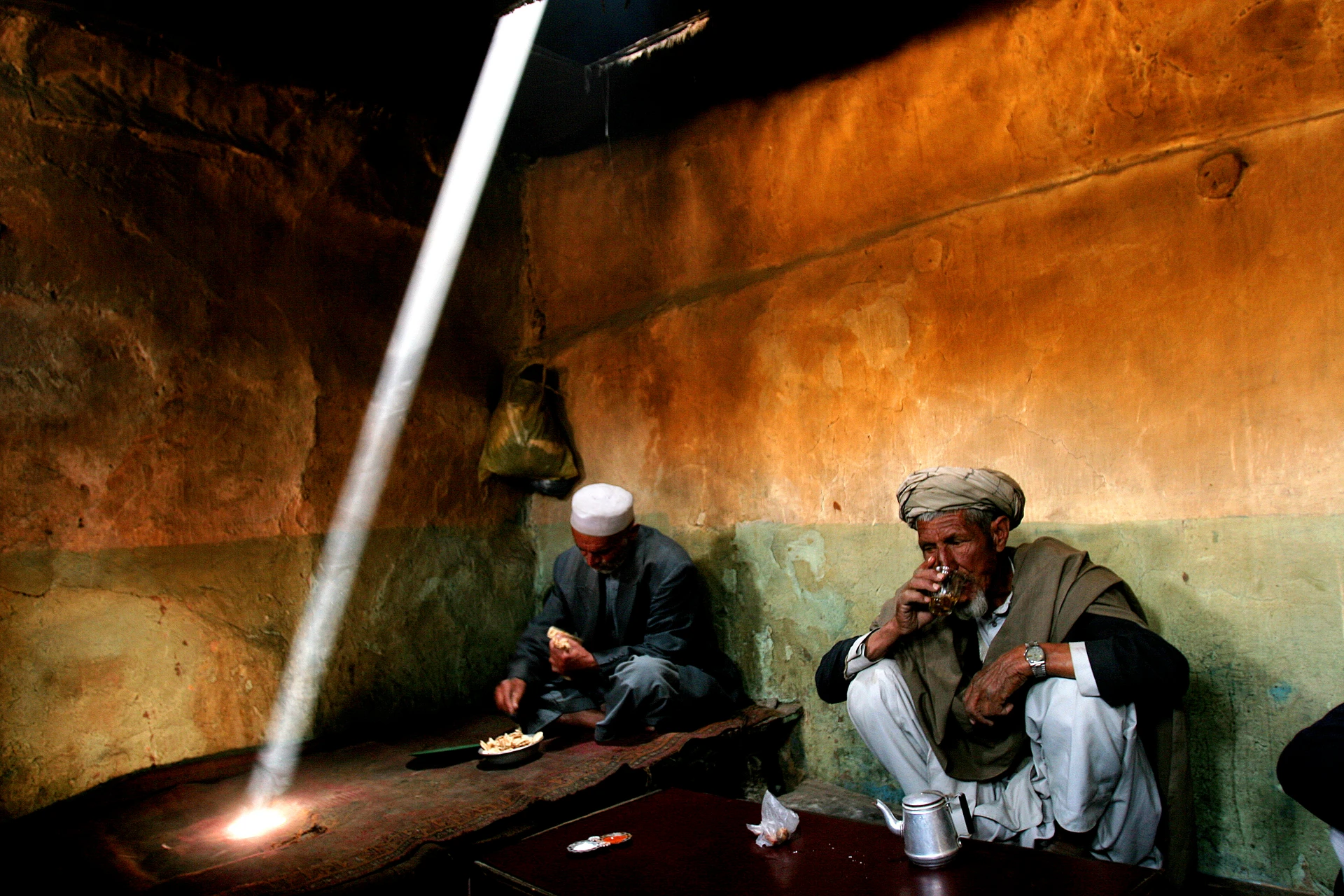 兩名戴阿富汗帽的男子坐在簡陋房間裡吃喝，一束陽光透過屋頂照在地毯上。