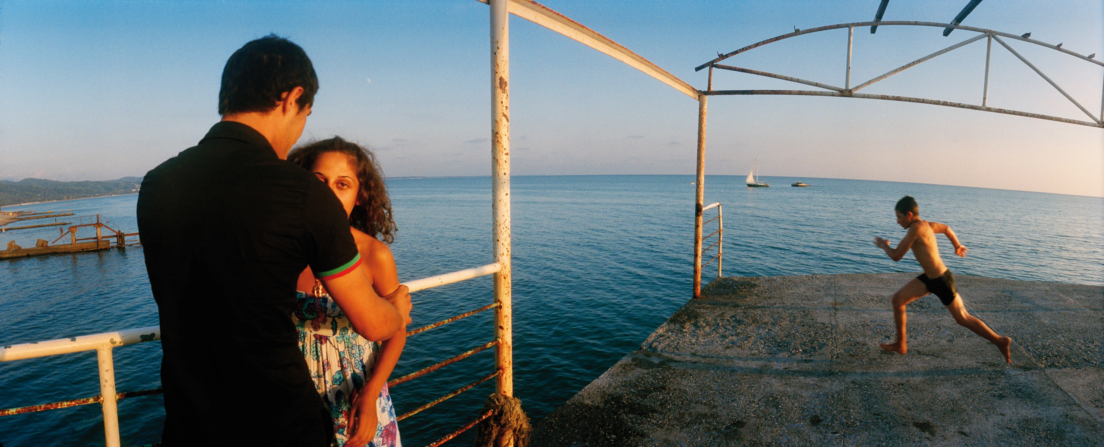 A child runs barefoot across a concrete platform over the sea on which a dilapidated metal railing structure stands. The child is about to jump into the water. In the foreground, a man holds a woman by her arms.