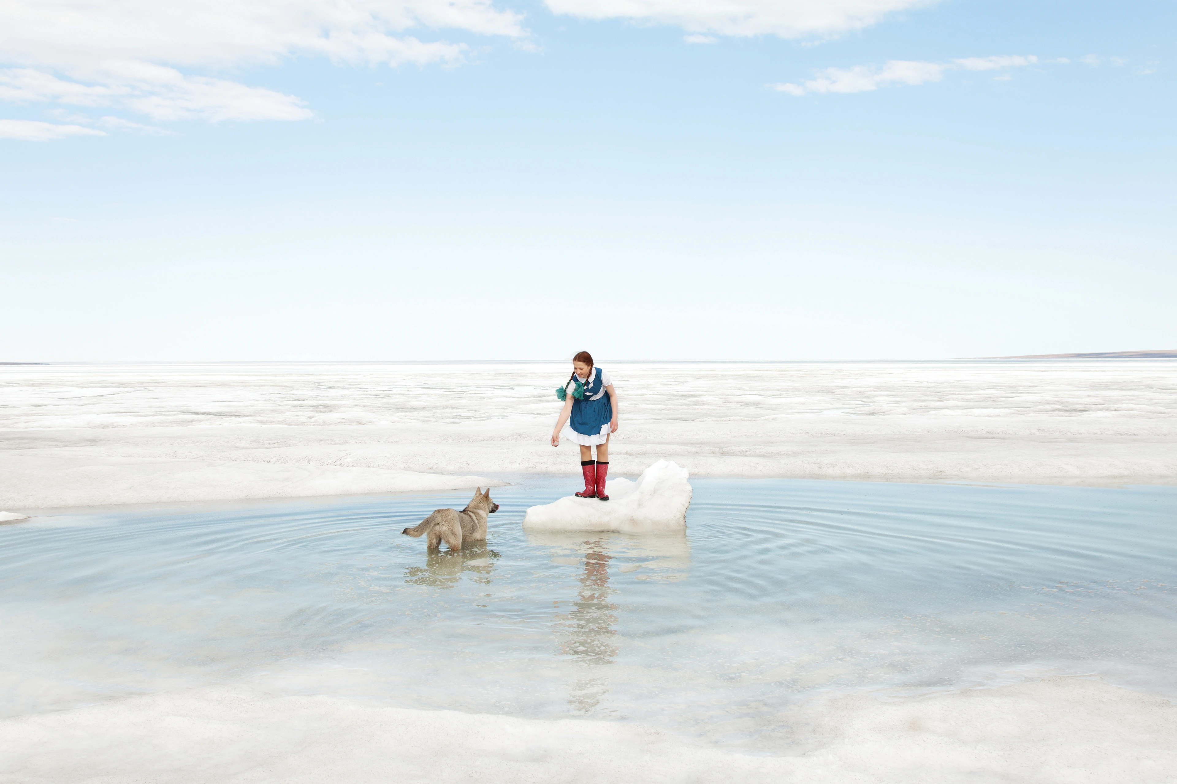 A girl stands in the centre of the picture on a small ice floe in shallow water. She calls and reaches out to a dog wading in the water next to her.