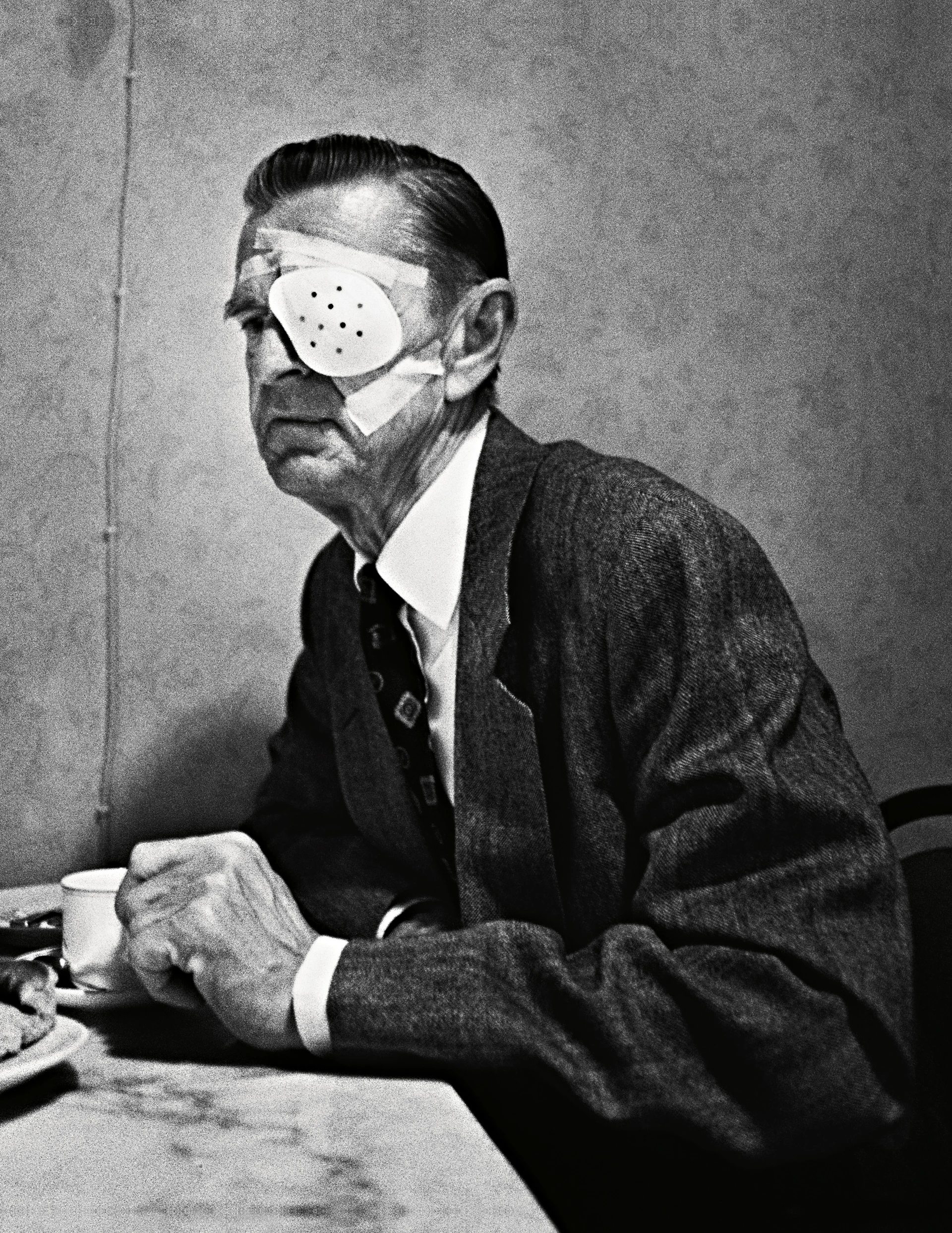 Black-and-white image: An elderly man in a suit sits at a table wearing a large light-coloured eye shield with plasters over his eyes.