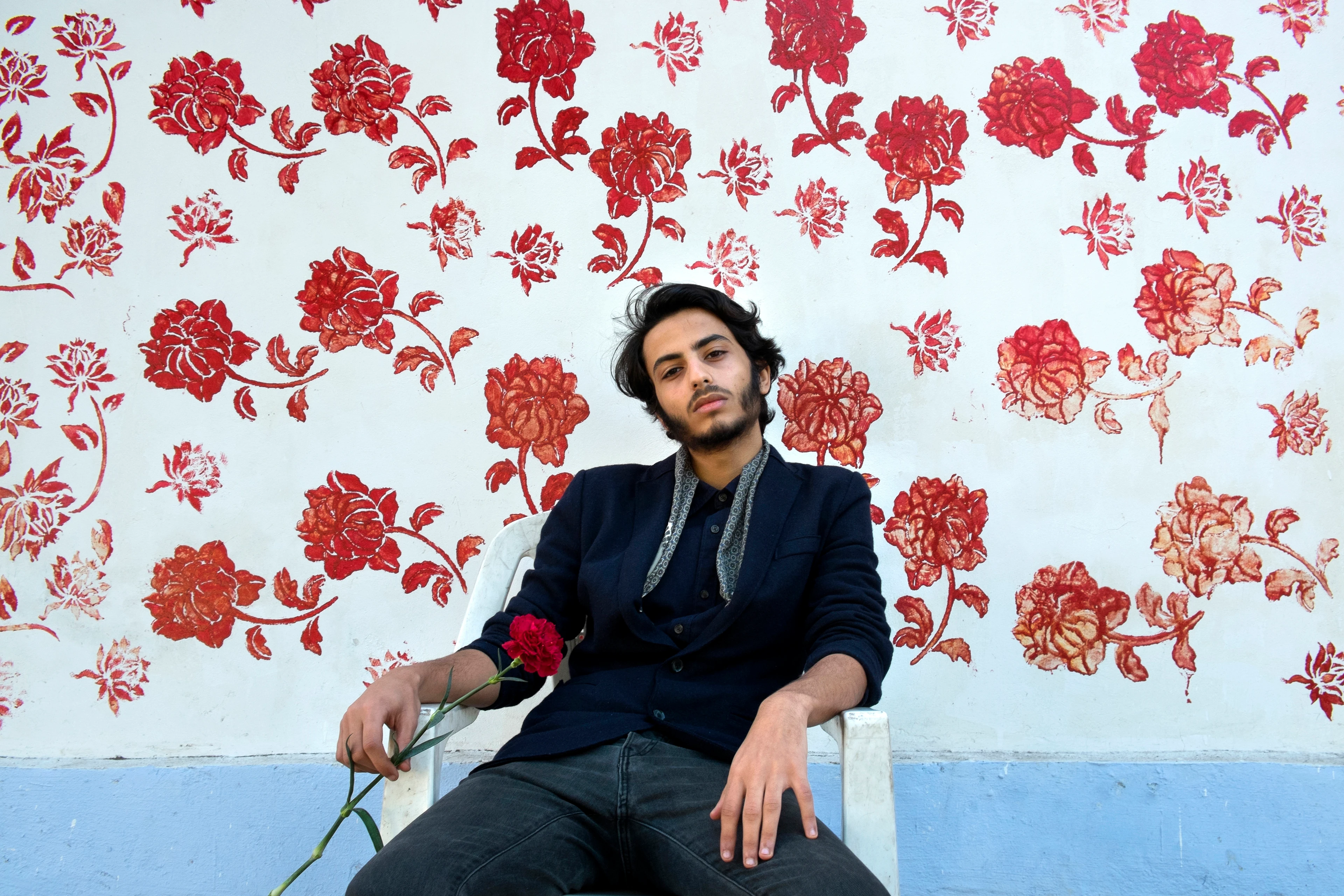 Portrait: A young man with a confident look sits relaxed on a white chair in front of a wall painted with red floral patterns, holding a rose.