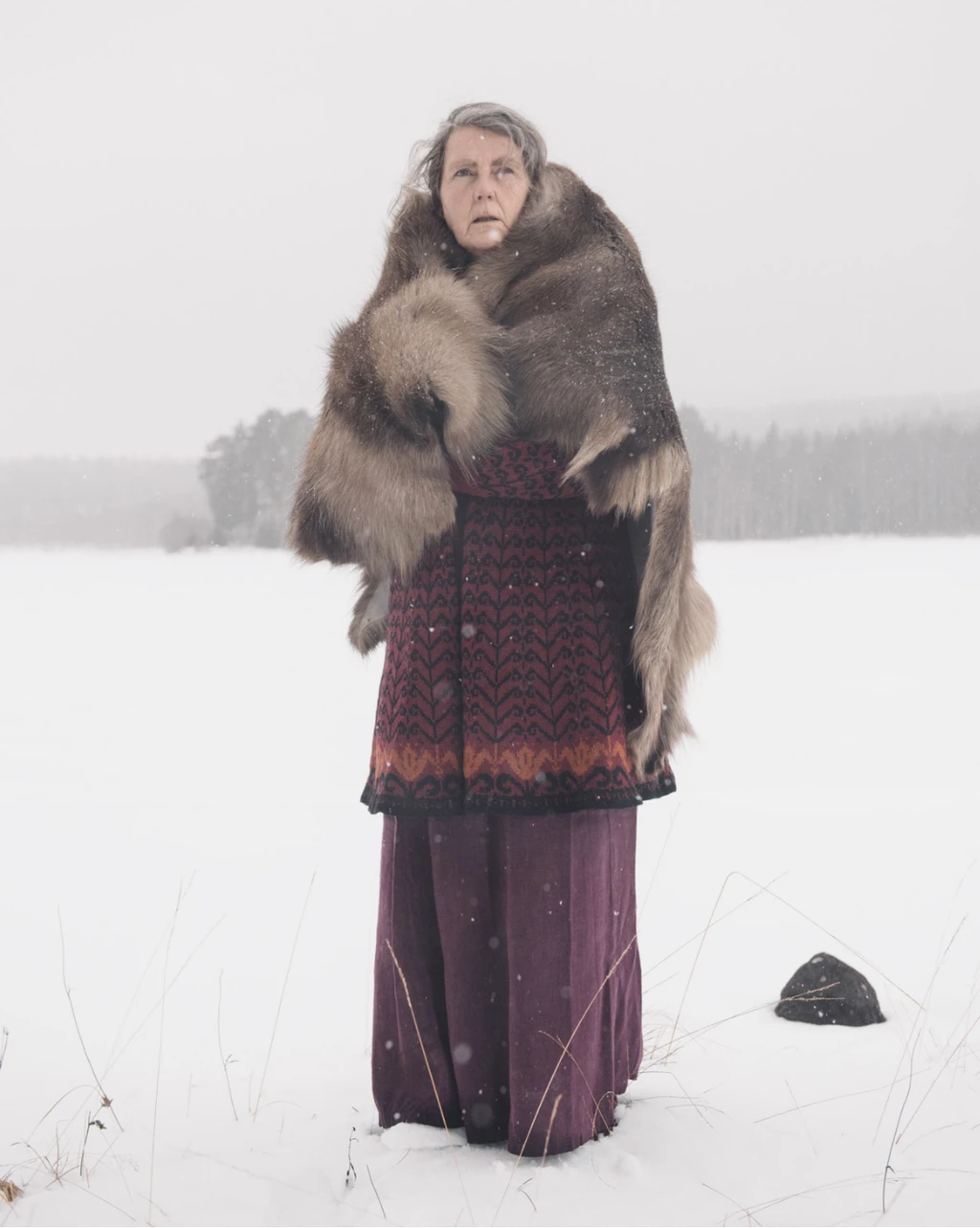 An elderly woman stands in the snowy outdoors, wearing a long dress and with a large pelt draped over her shoulders.