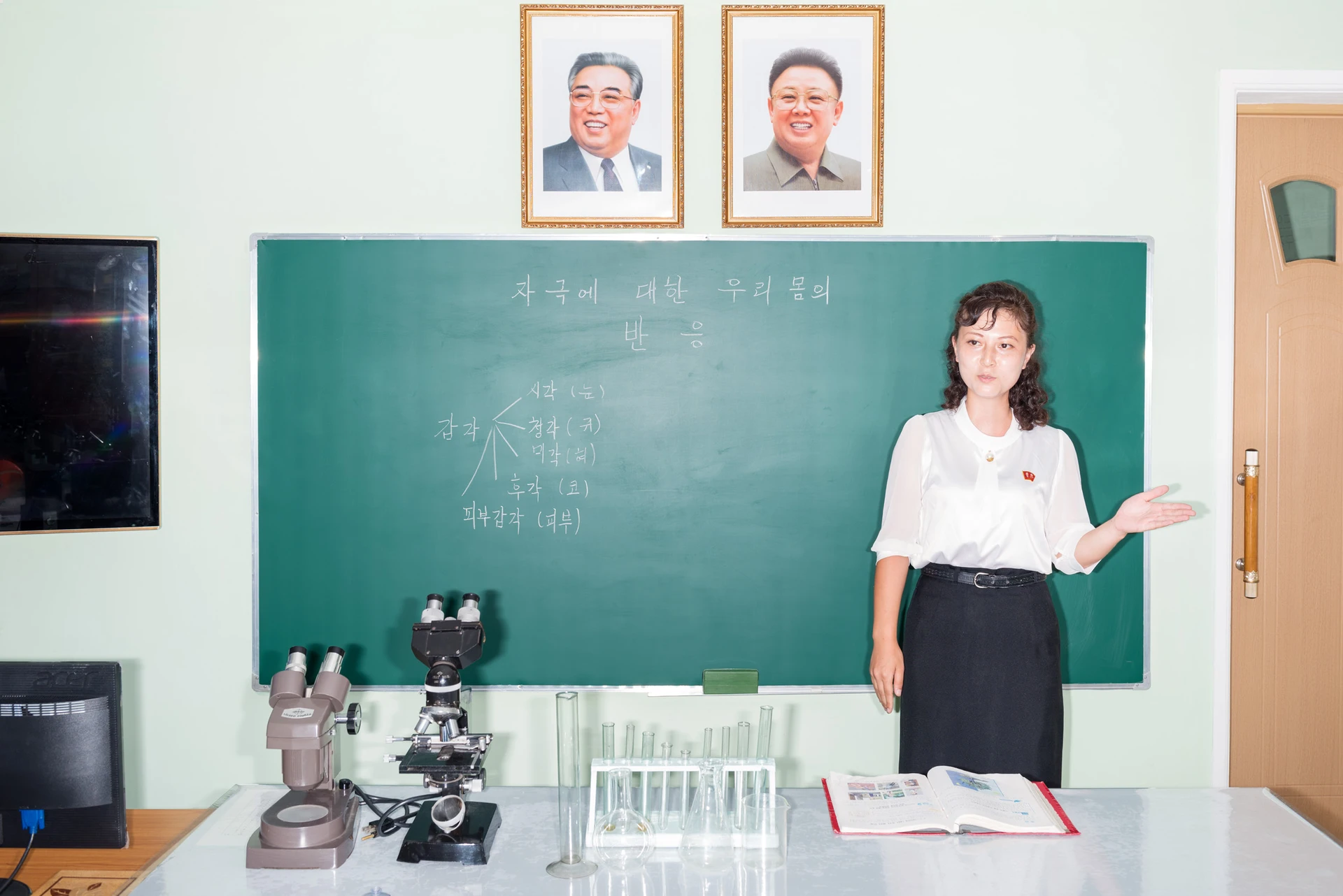 A teacher wearing a white blouse stands in front of a green blackboard, giving a lecture. Microscopes and test tubes are placed on a table in front of her, and two portraits of smiling North Korean political leaders hang above the blackboard.