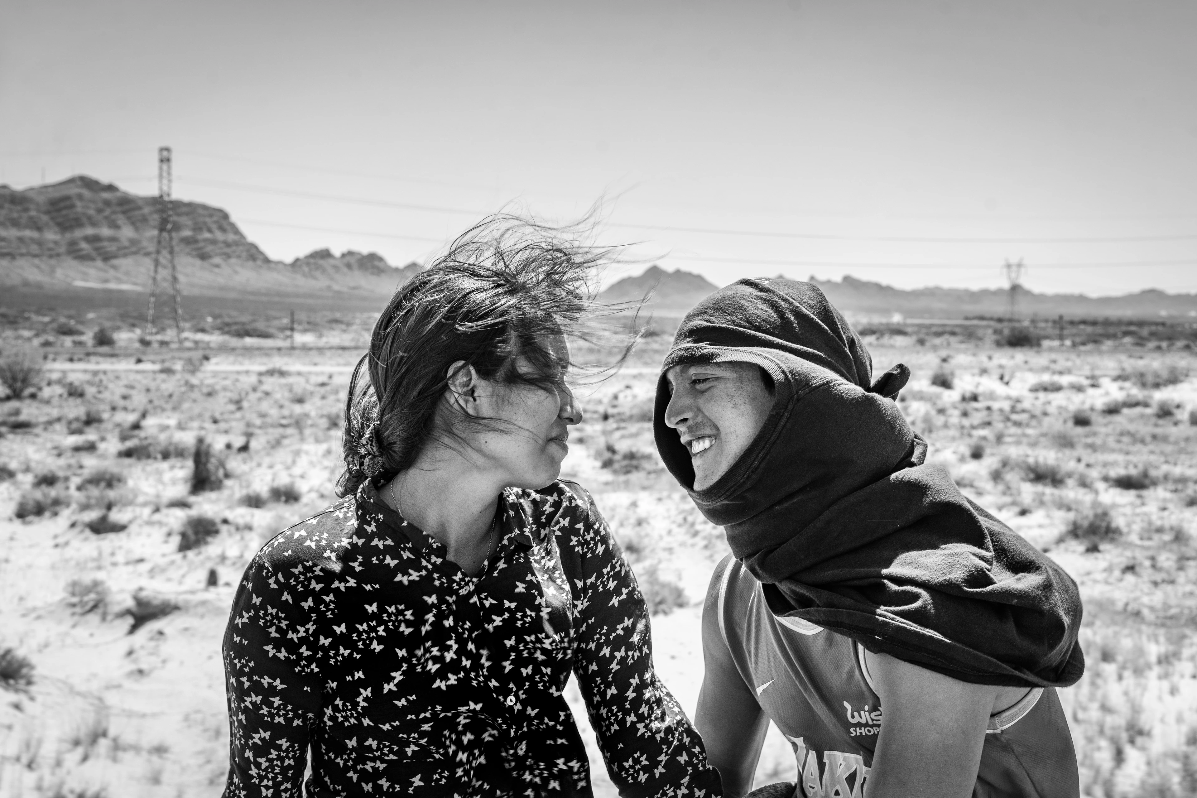 Black-and-white image: A young couple look into each other’s eyes, smiling and in love. A desert landscape stretches out behind them, her hair and his scarf blowing strongly in the wind.