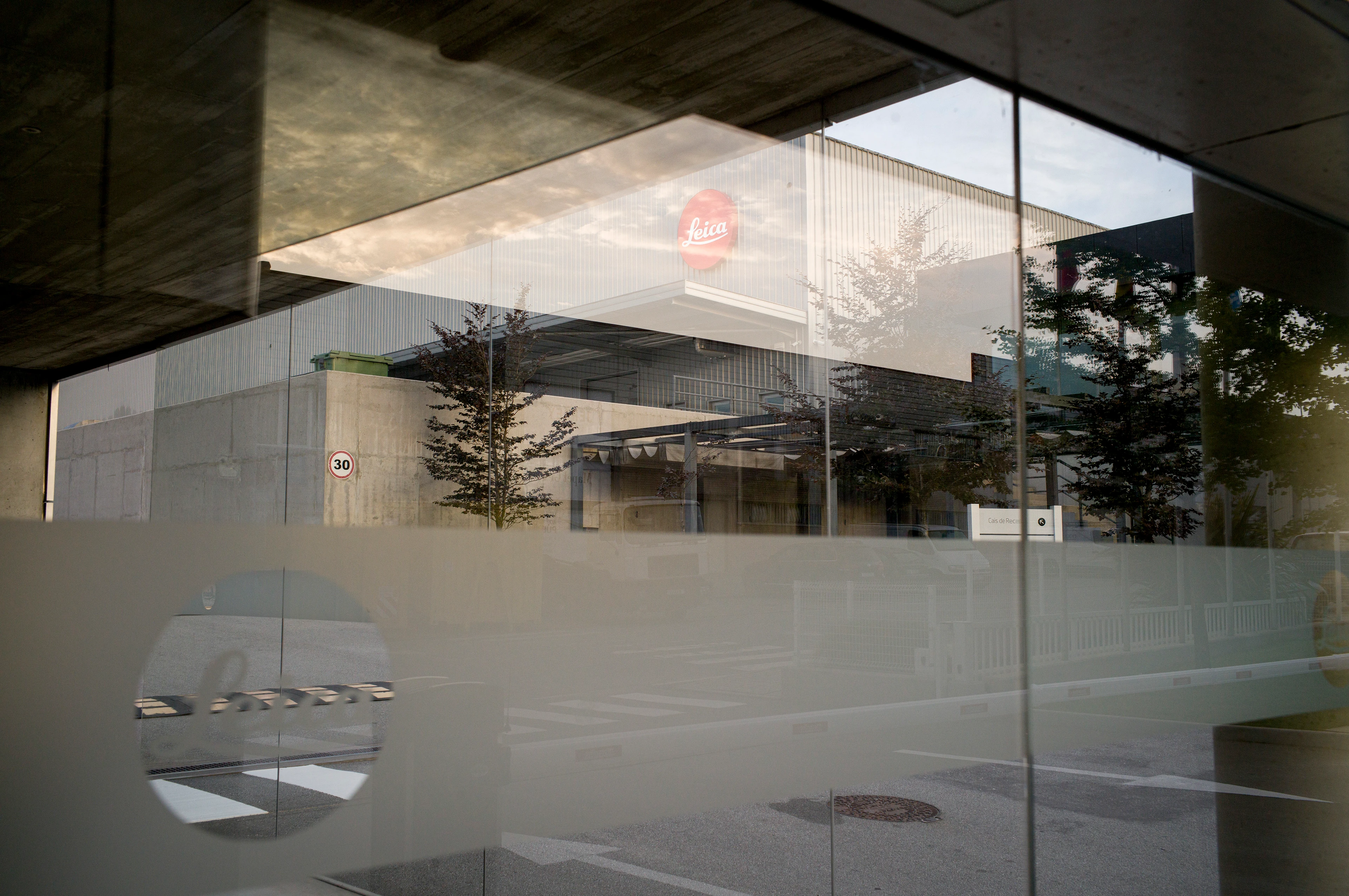 Außenaufnahme vom Eingang eines modernen Fabrikgebäudes mit großen Glasflächen; die Spiegelung zeigt Bäume, Himmel und das rote Leica Logo auf der Fassade.
