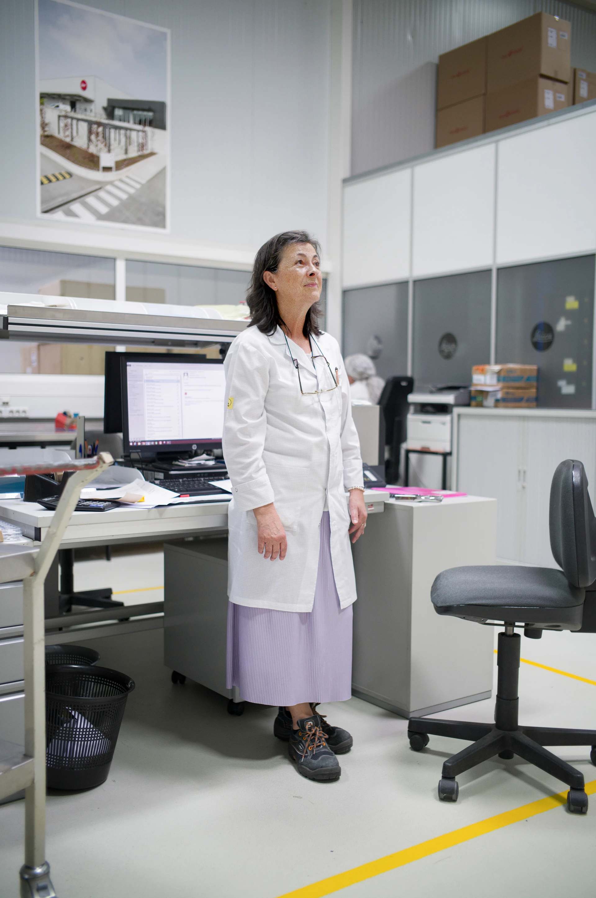 Eine ältere Frau im weißen Laborkittel und lila Rock posiert stehend und freundlich zur Seite blickend in einem Büro-Laborraum neben einem Schreibtisch mit mehreren Monitoren und Laborunterlagen.