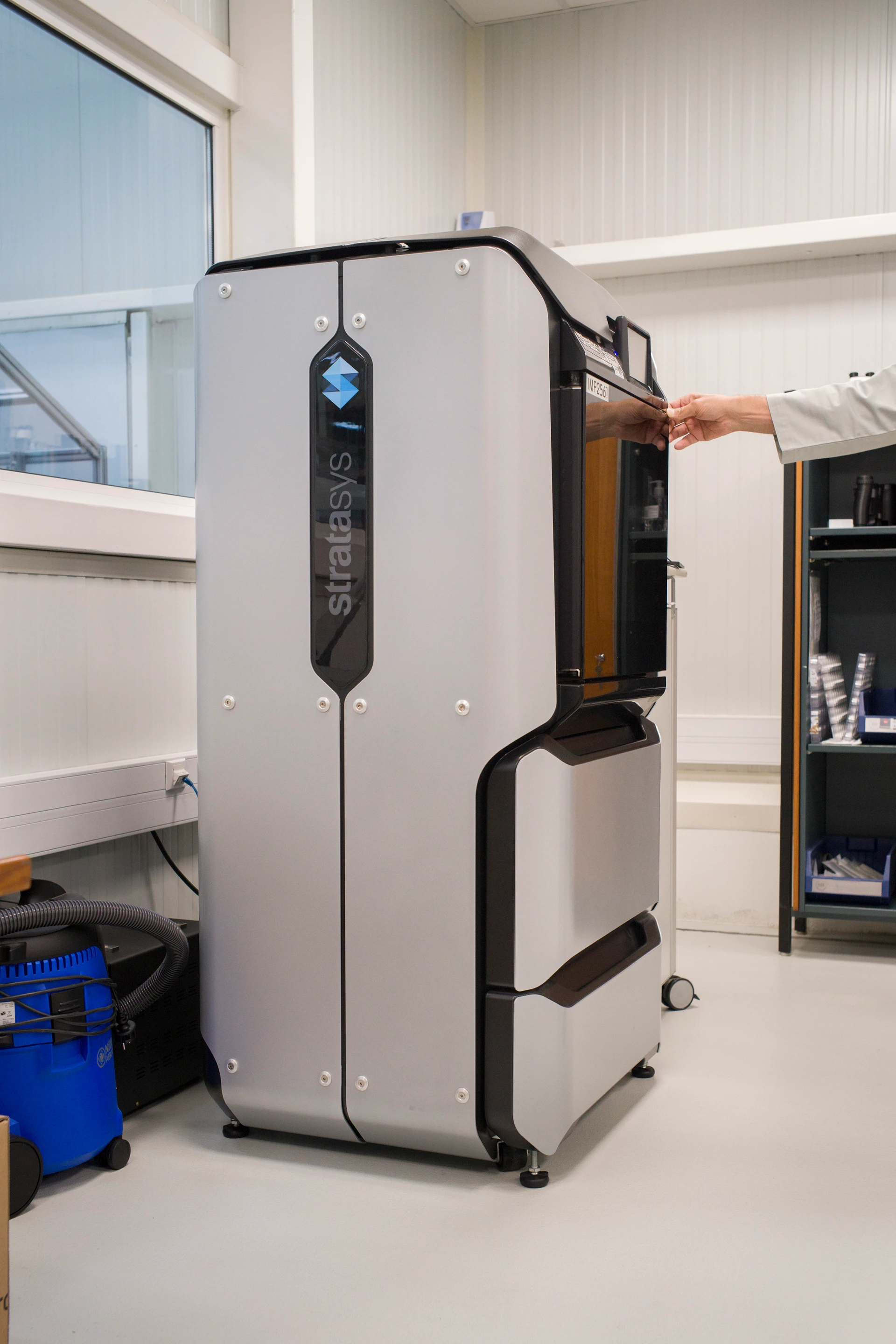 Ein großer industrieller 3D-Drucker in einer sauberen Laborumgebung; ein einzelner Arm in Laborkittel kommt von der Seite ins Bild und bedient das Gerät.