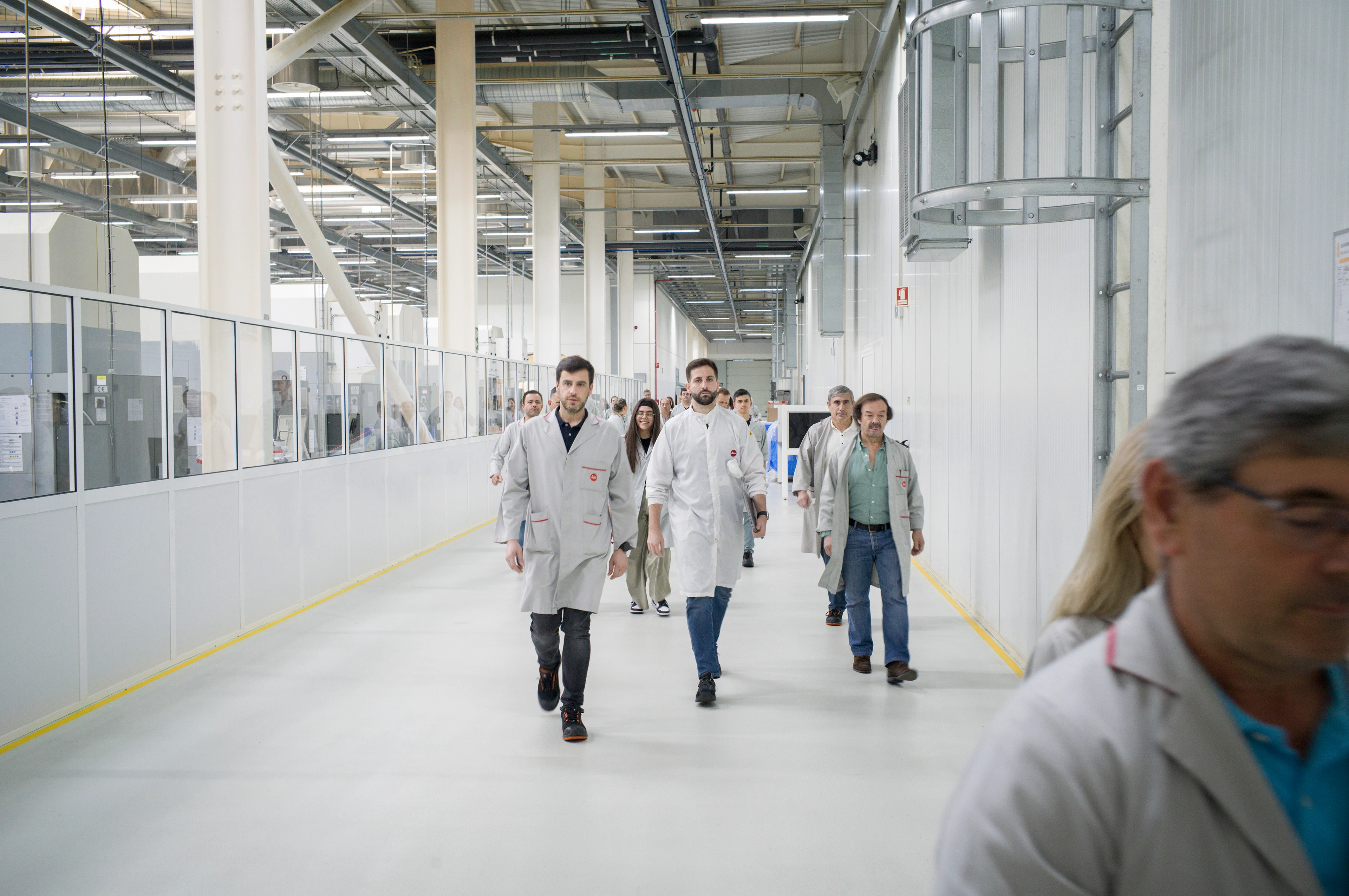 Eine Gruppe von Personen in weißen Laborkitteln geht durch einen breiten, hellen Industrie- oder Forschungsflur auf die Kamera zu; Leitungen und Metallkonstruktionen verlaufen über ihnen an der Decke.