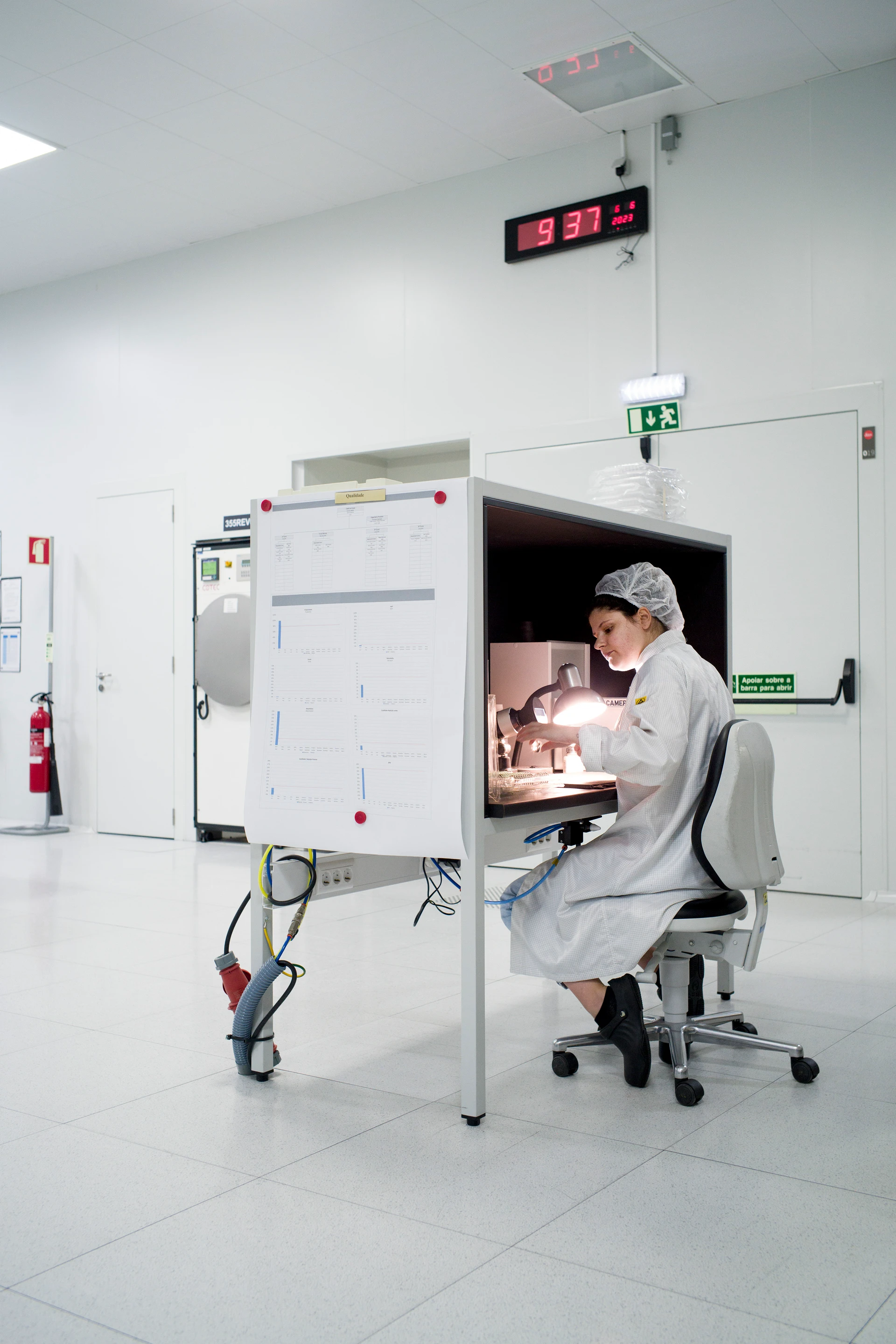 Eine Mitarbeiterin im weißen Labor­kittel und Haarnetz sitzt an einem Arbeitsplatz in einem Reinraum und untersucht ein Glasteil unter einer starken Lampe; über ihr hängt eine digitale Uhr an der Wand.