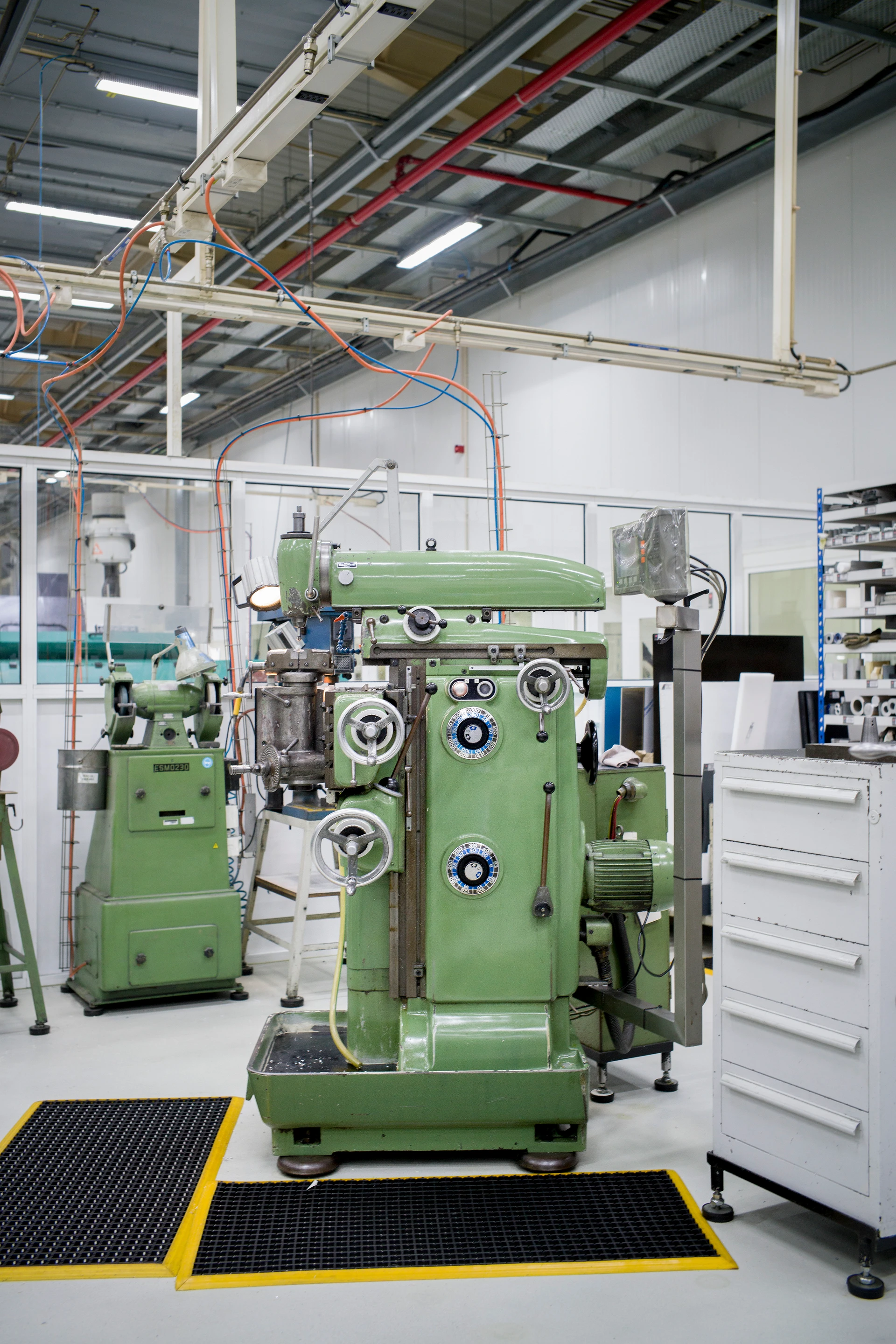 Große, industrielle grüne Maschine mit mehreren Knäufen und Anzeigen in einer Produktionshalle; Kabel und weitere grüne Geräte sind im Hintergrund sichtbar.