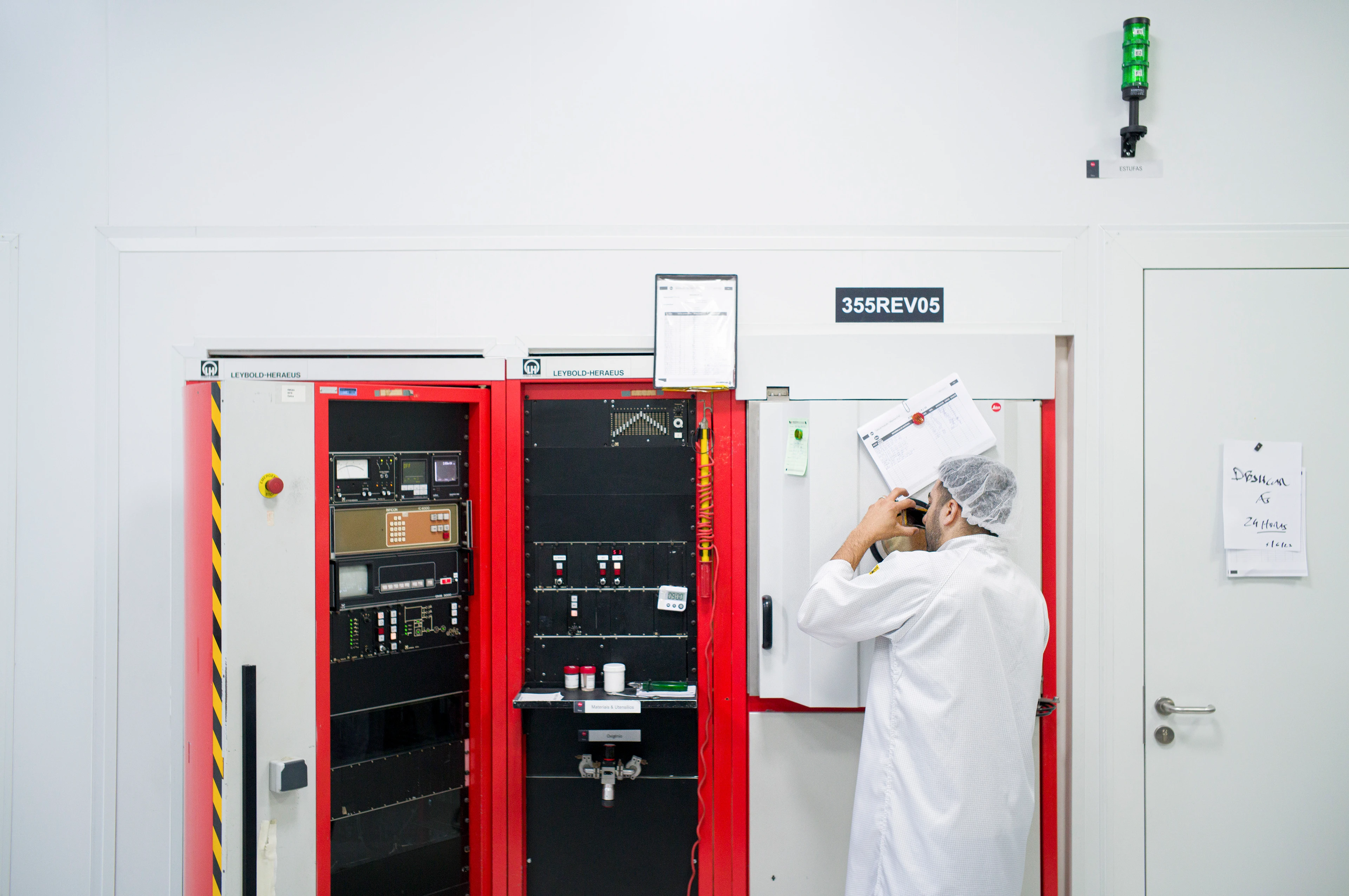Ein Techniker im weißen Labor­kittel und Haarnetz prüft Dokumente vor einem großen Schaltschrank mit rot-schwarzer Front und Anzeigen; ein grünes Warnlicht leuchtet über dem System.