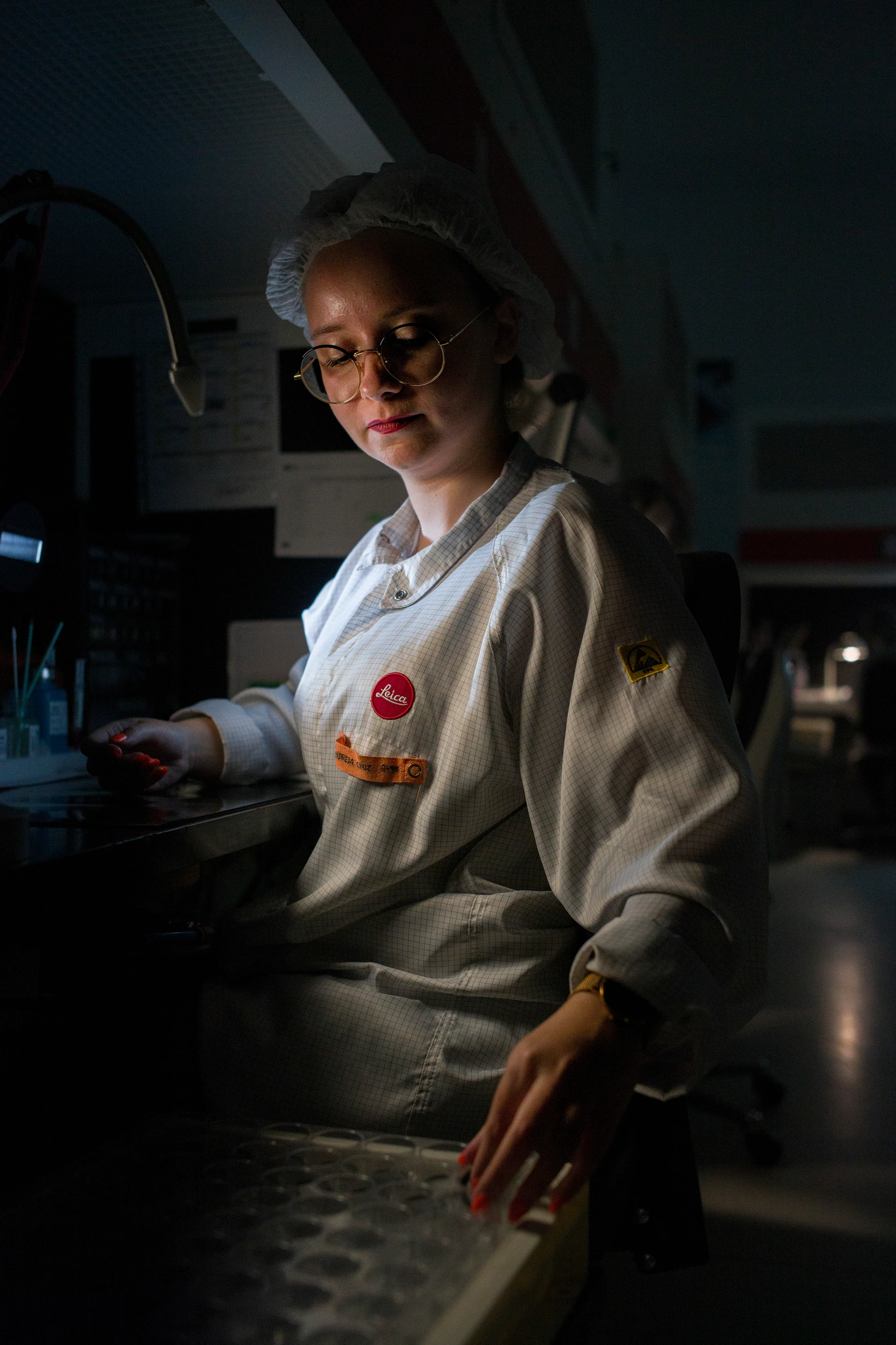 Eine junge Frau im weißen Leica Laborkittel und Haarnetz arbeitet konzentriert an einem Labortisch; sie trägt Schutzbrille und wird von einer einzelnen Arbeitsplatzlampe beleuchtet, während der Hintergrund dunkel bleibt.