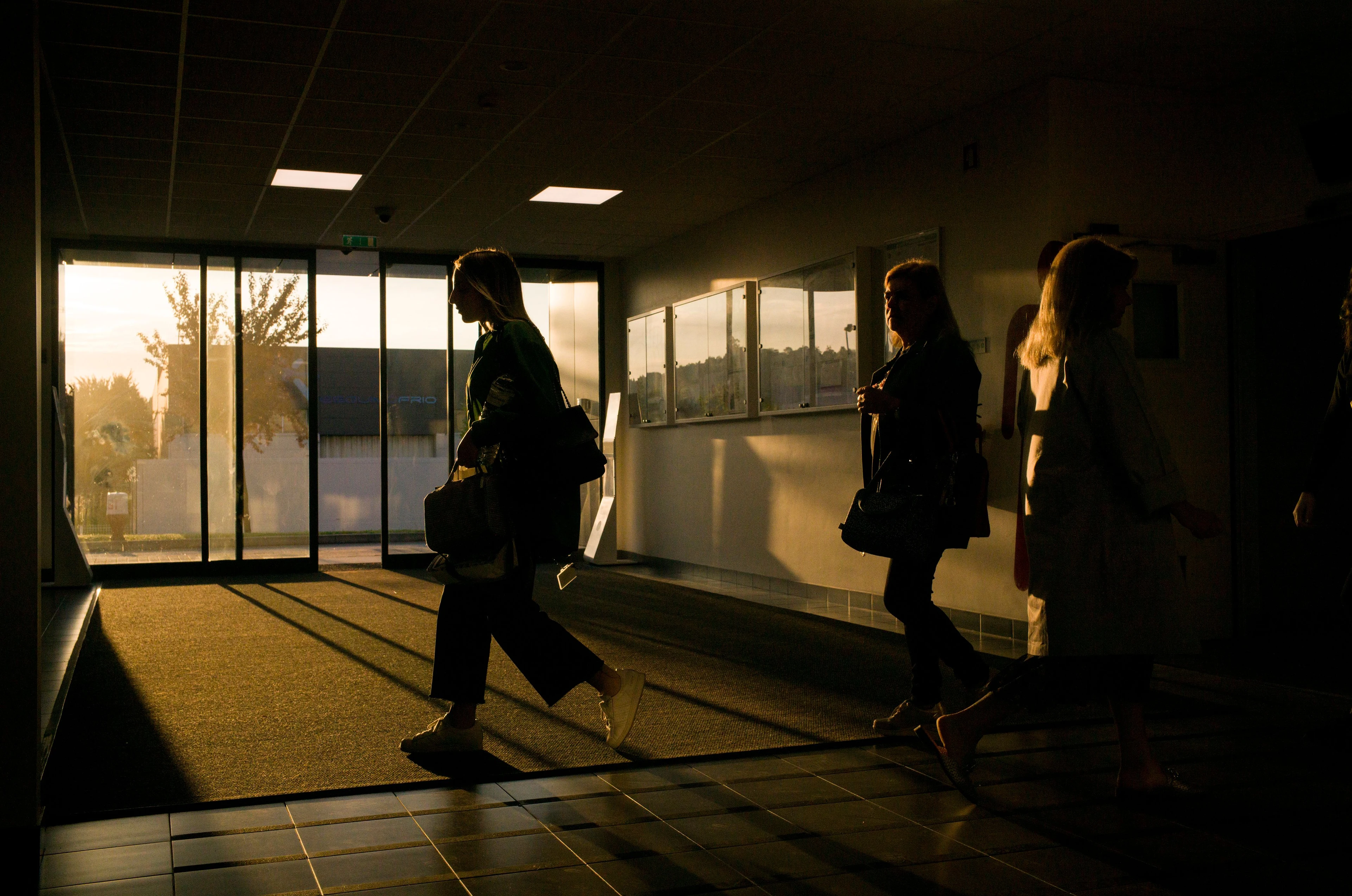 Mehrere Personen gehen in einem Firmenfoyer im Gegenlicht an großen Glas-­Eingangstüren vorbei; das warme Sonnenlicht erzeugt lange Schatten auf dem Boden und wirkt wie ein Schichtwechsel am Ende des Arbeitstages.
