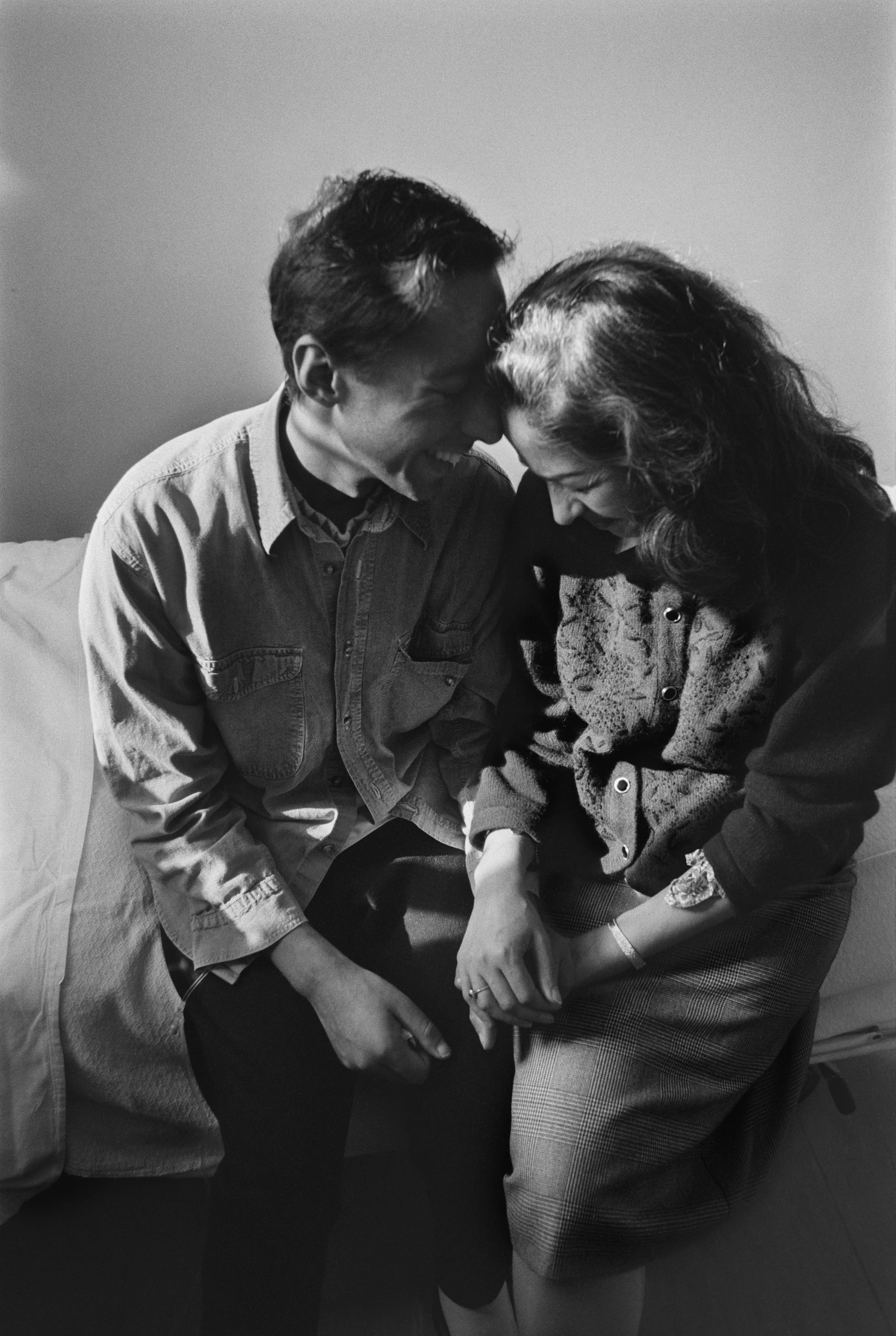 Ritratto suggestivo in bianco e nero di una coppia che, testa contro testa, è seduta su un letto d’ospedale nel reparto AIDS e sorride.