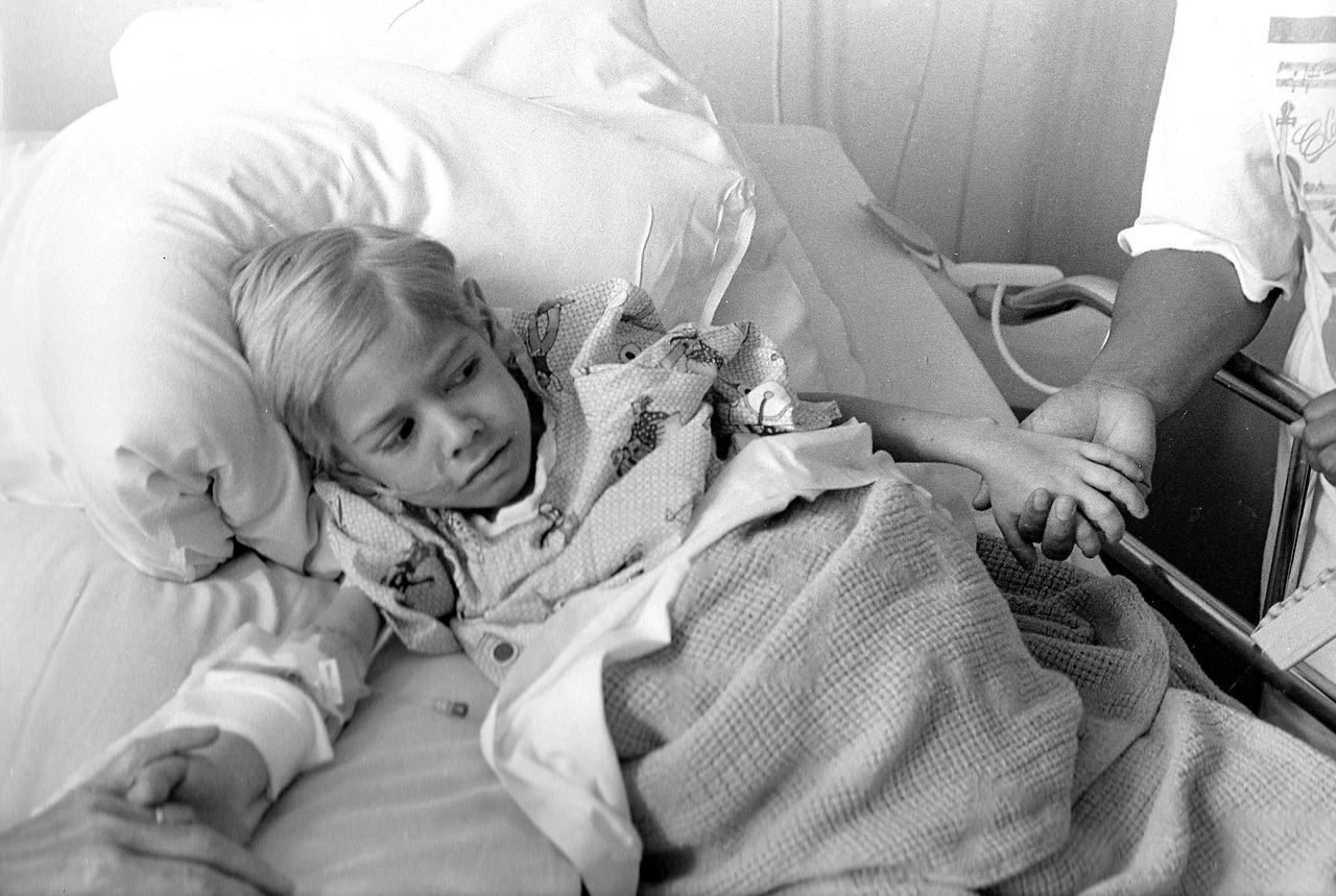 Foto in bianco e nero di un bimbo malato di AIDS in un letto d’ospedale mentre due adulti gli tengono le mani.