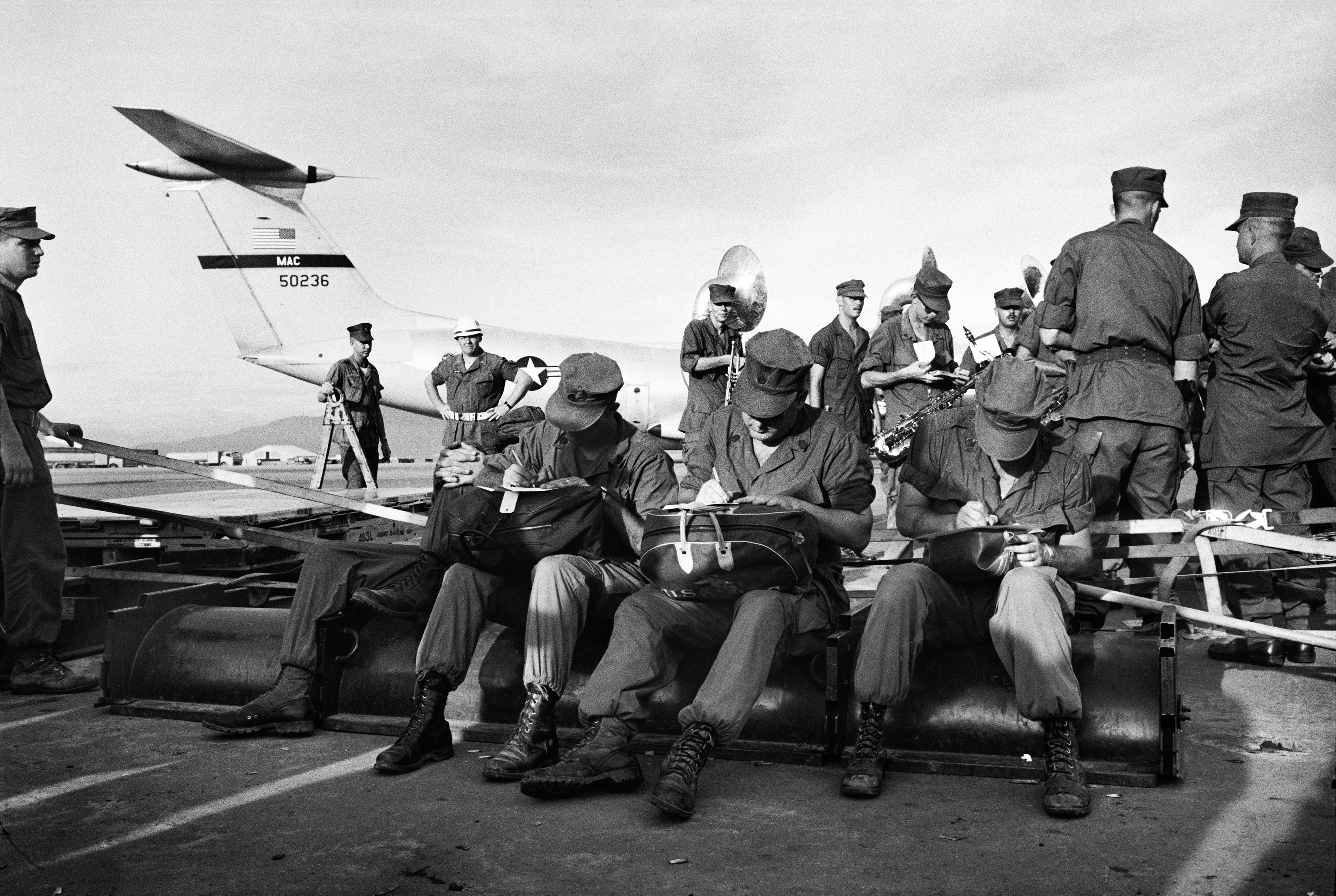 Motyw czarno-biały, wojna w Wietnamie, żołnierze czekają na pasie startowym, w tle inni żołnierze z instrumentami muzycznymi