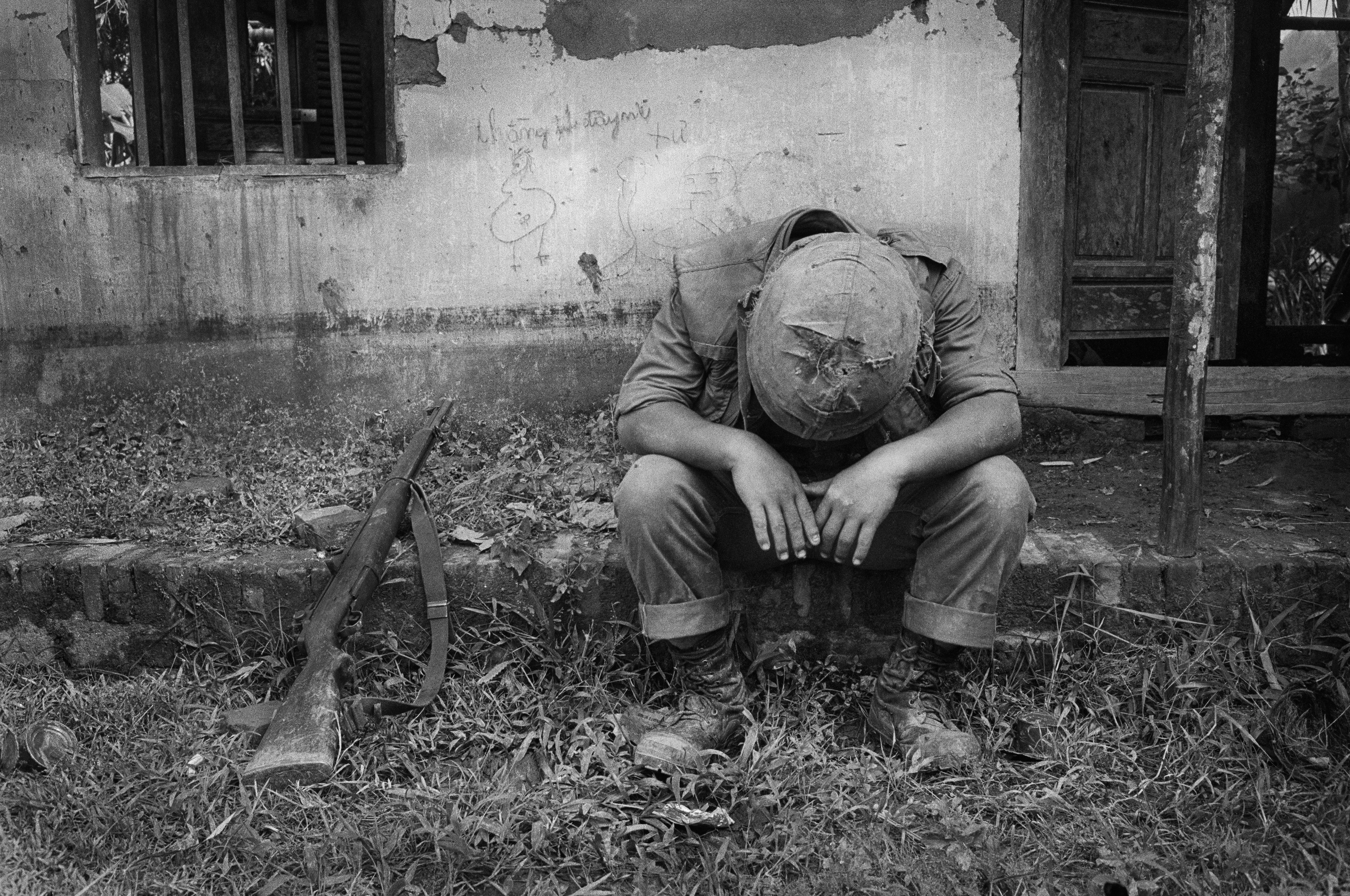 Motyw czarno-biały, wojna w Wietnamie, wyczerpany żołnierz siedzi przed ścianą domu, z opuszczoną głową, karabin leży obok niego