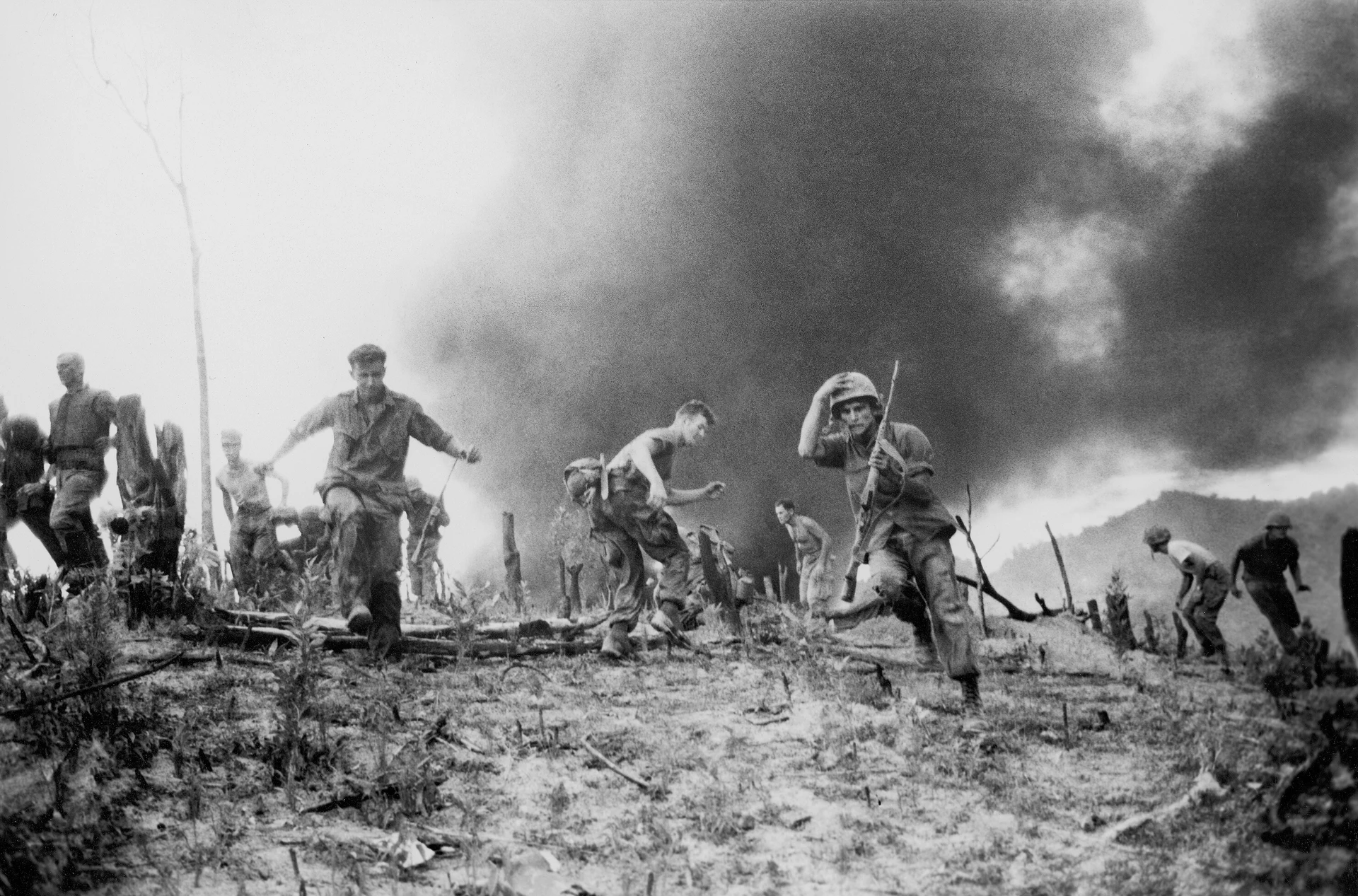 Motyw czarno-biały, wojna w Wietnamie, żołnierze biegną przez pole, chmury dymu na horyzoncie