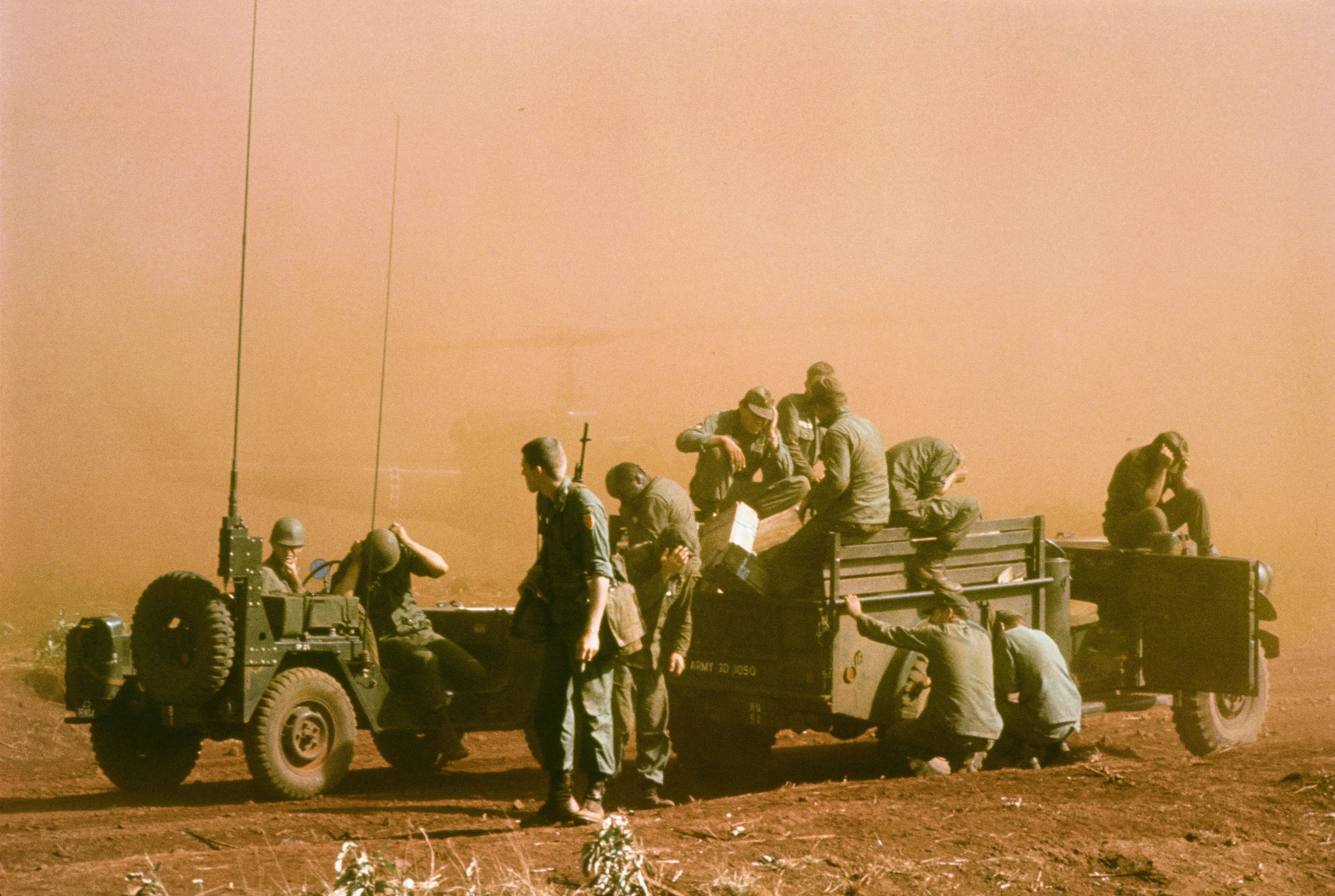 Wojna w Wietnamie, kilku żołnierzy chroni twarz przed chmurą pyłu, jeden z dwóch samochodów terenowych ma antenę radiową, w drugim naprawiane jest tylne koło 