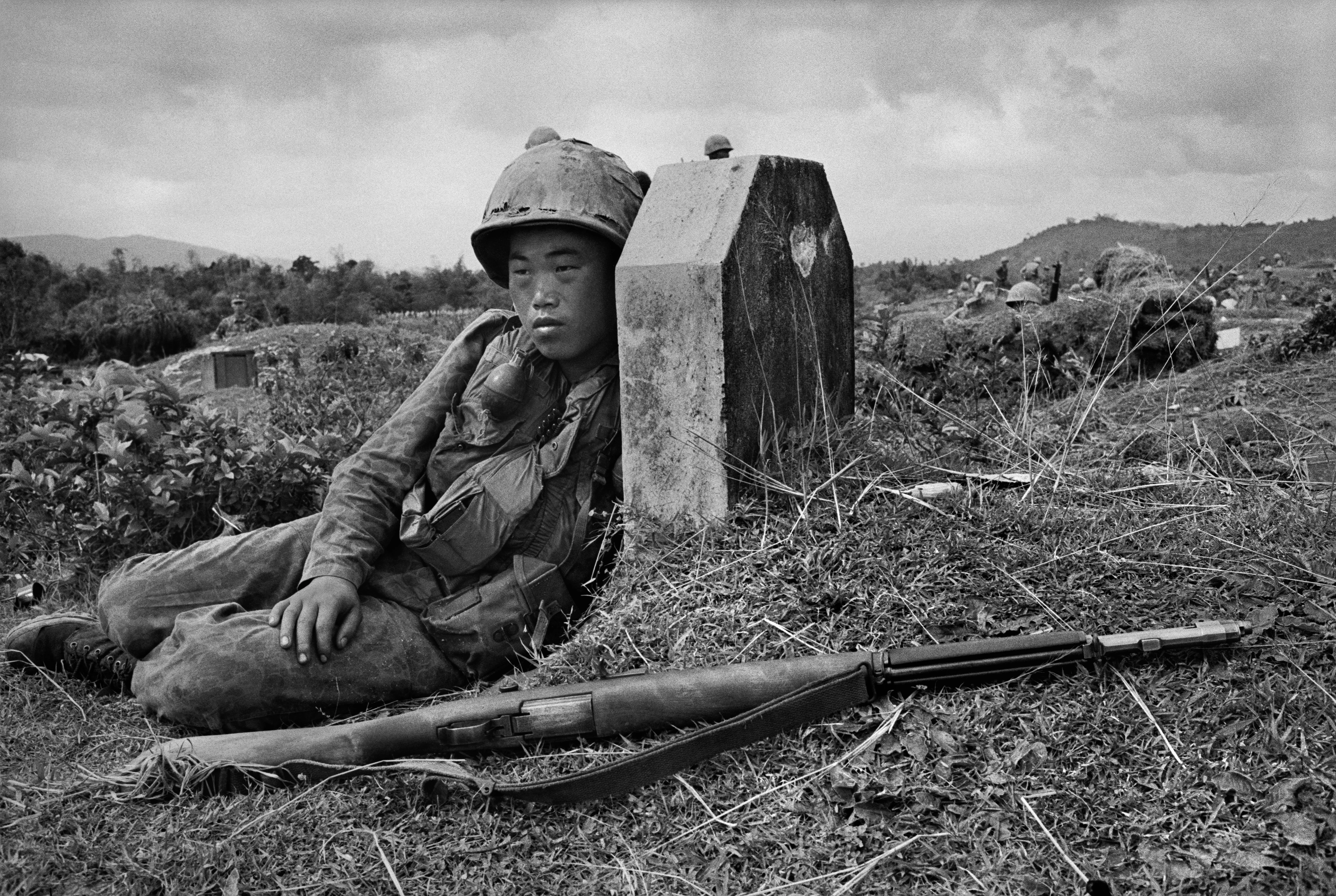 Motyw czarno-biały, wojna w Wietnamie, żołnierz kuca przy nagrobku, w tle inni żołnierze