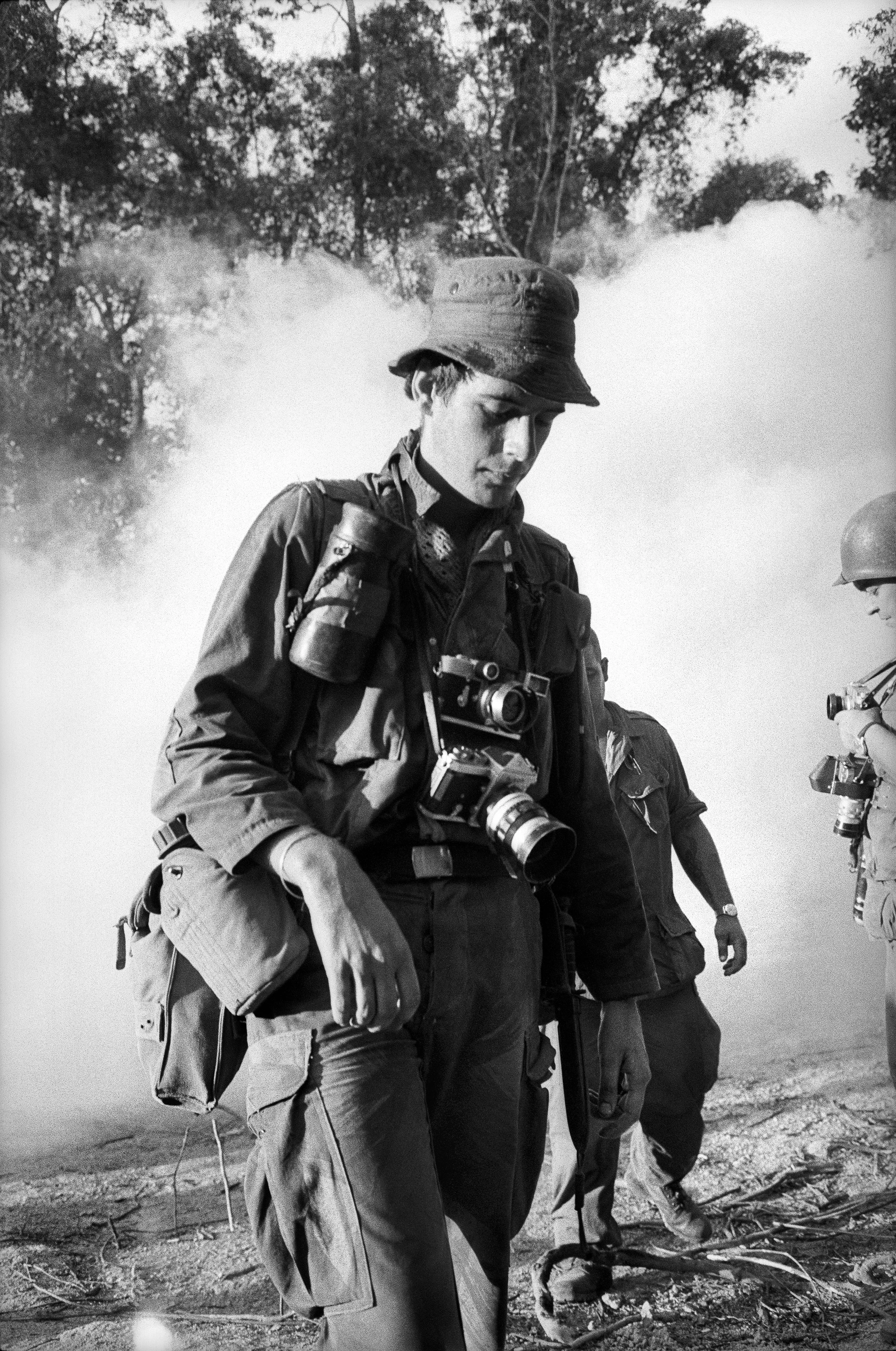Black-and-white portrait of Tim Page during the Vietnam War loaded down with camera equipment