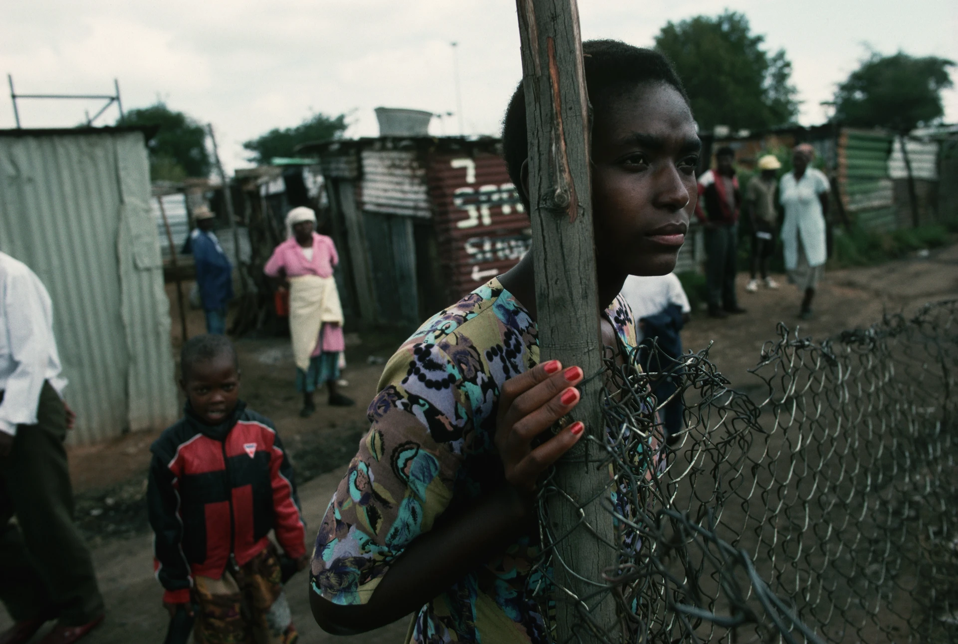 Una donna di colore sudafricana, con le unghie dipinte di rosso, è in piedi, pensierosa, appoggiata al palo di legno di una recinzione e guarda in lontananza; dietro di lei ci sono diversi abitanti di un insediamento, anziani e giovani.