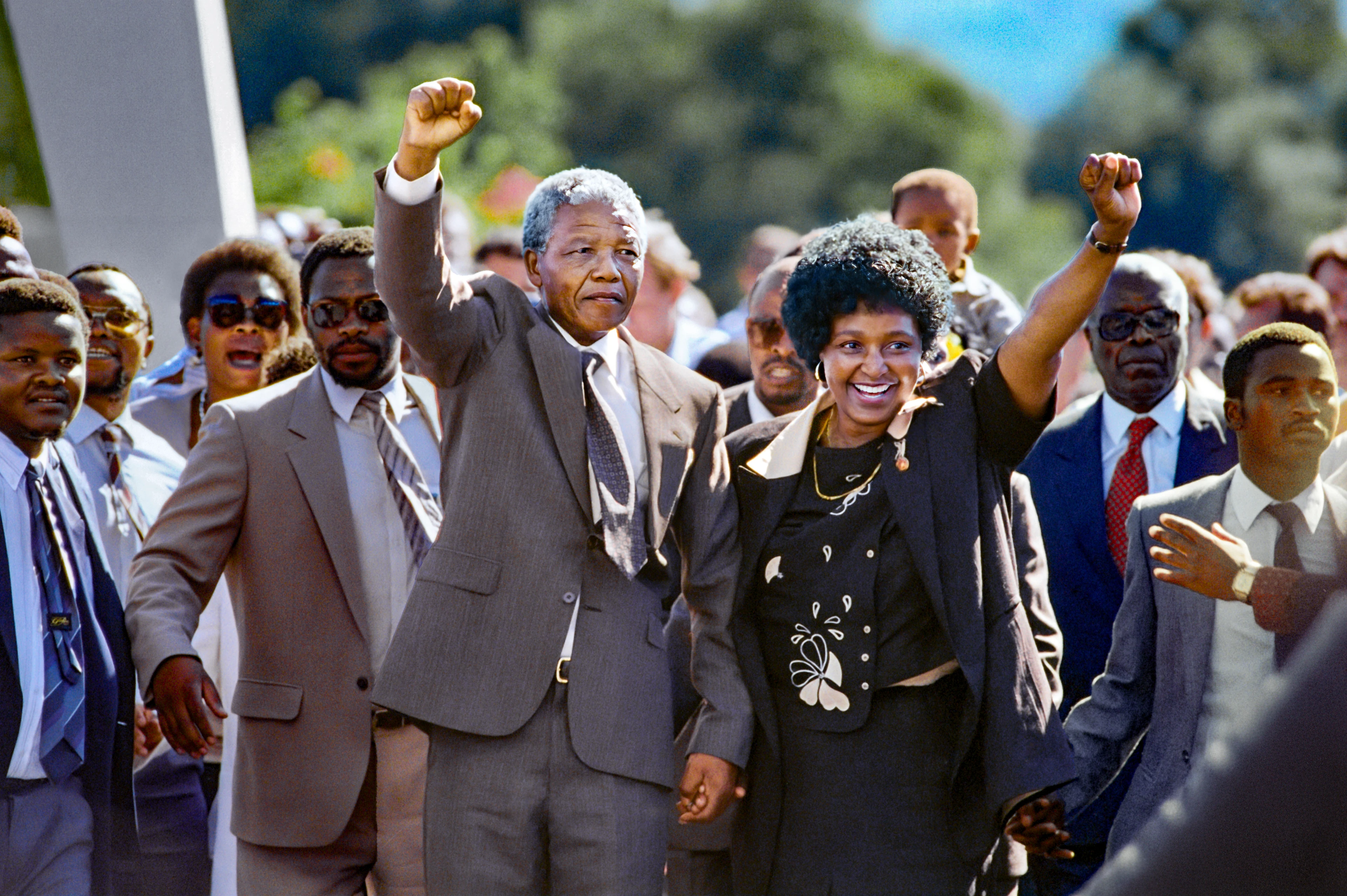 In una giornata di sole, Nelson Mandela e sua moglie alzano il pugno combattivi, dietro di loro un intero seguito di uomini in giacca e cravatta.
