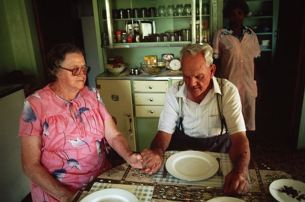 Una coppia di anziani bianchi si tiene per mano mentre prega seduta al tavolo da pranzo, sullo sfondo una donna di servizio di colore che aspetta paziente.