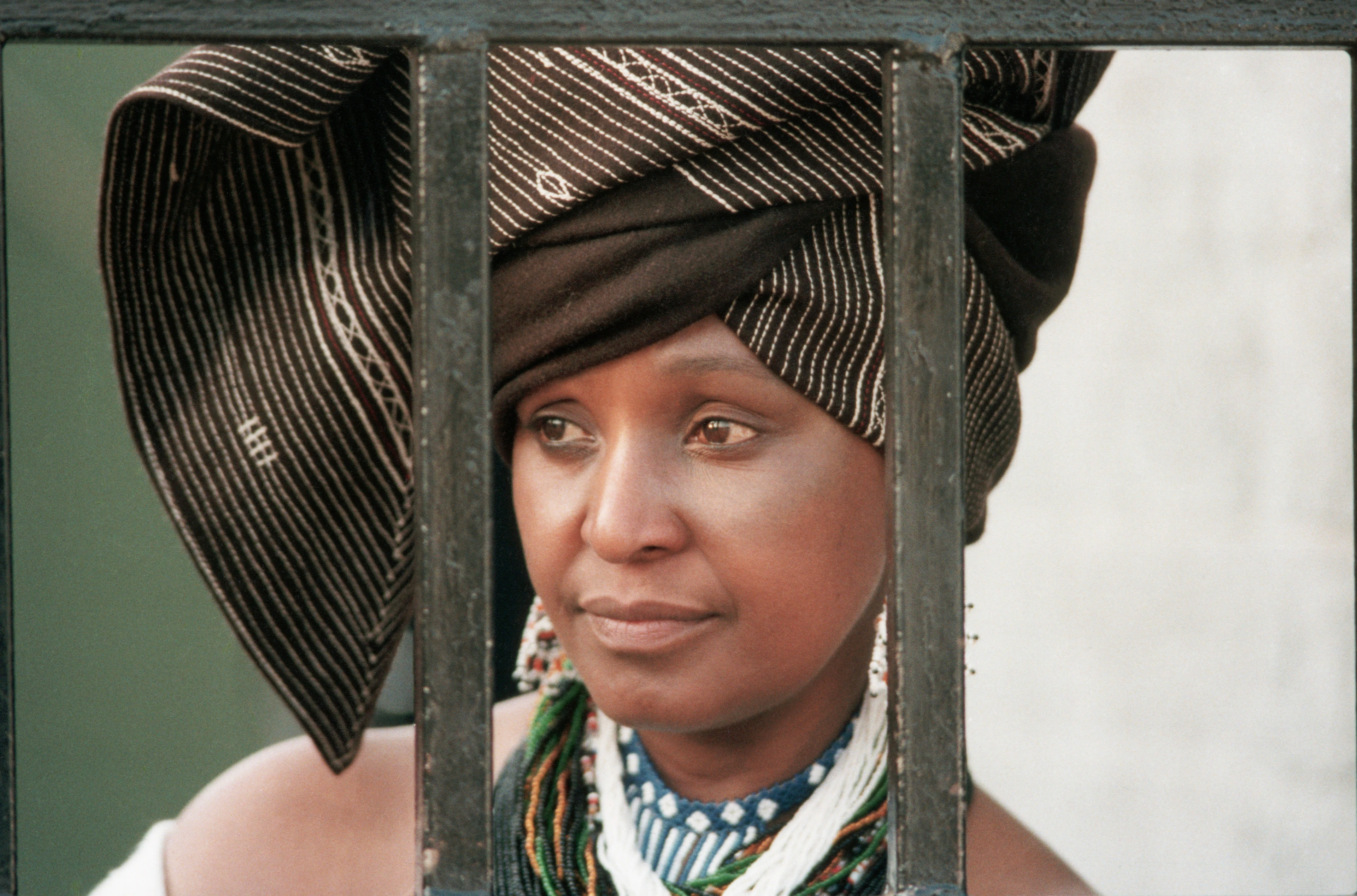 Winnie Mandela con un vistoso copricapo elegante e una collana dietro le sbarre di un cancello che guarda di lato pensierosa.