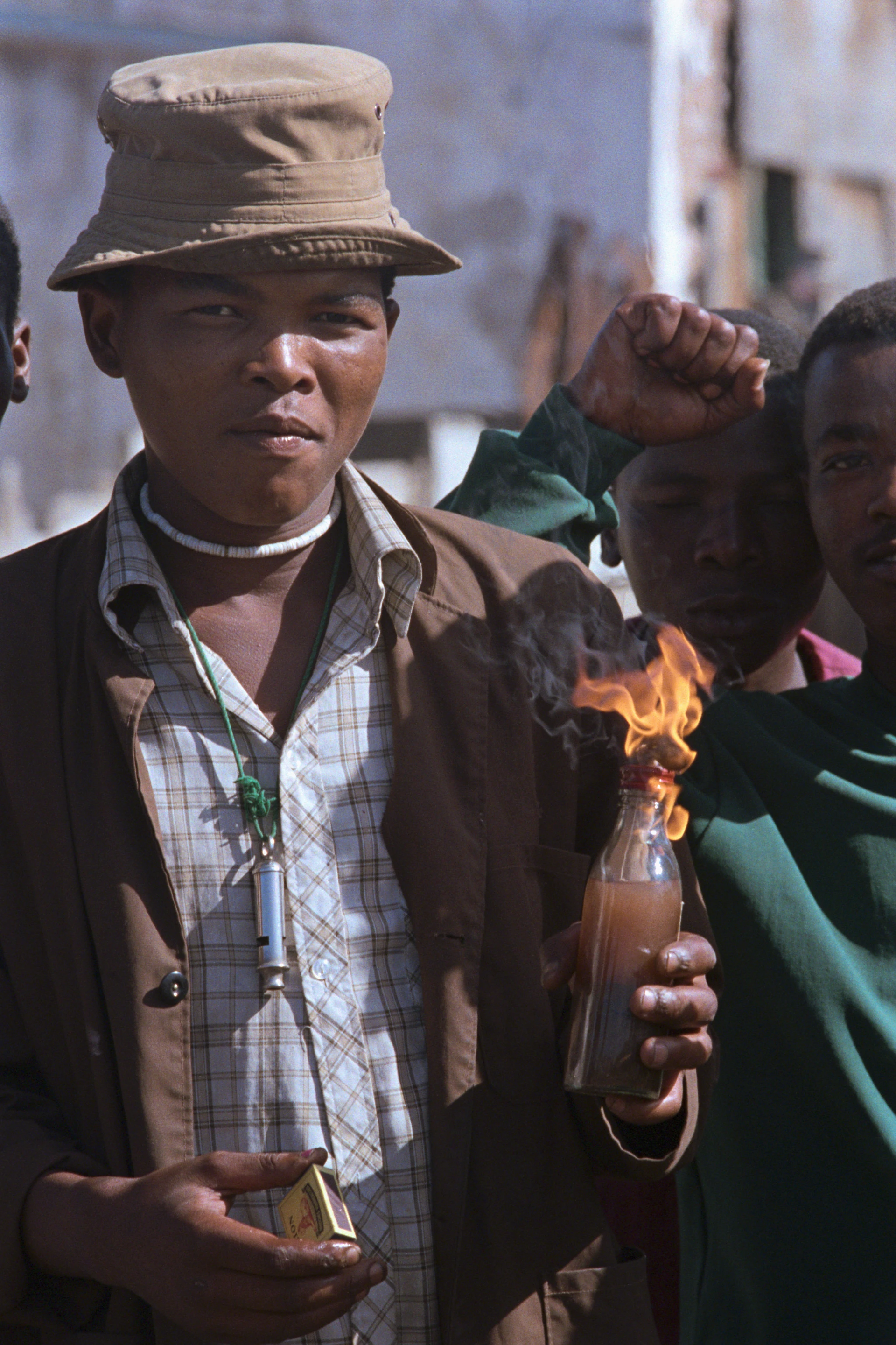 Un giovane di colore con cappello tiene una bottiglia molotov accesa nel mezzo di una protesta e guarda verso la fotocamera con espressione determinata.