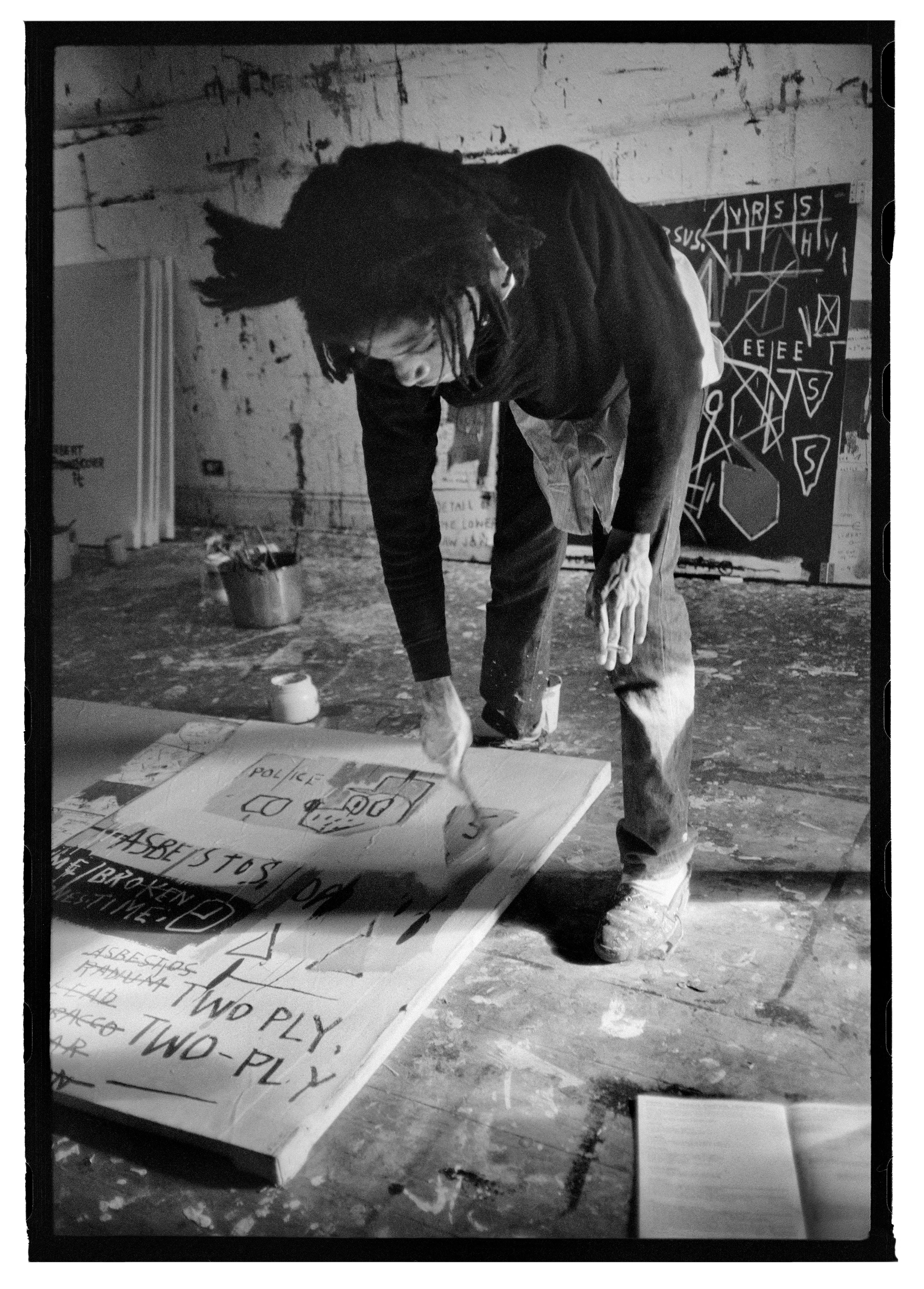 Fotografía en blanco y negro de Jean-Michel Basquiat, de pie inclinado hacia delante mientras pinta un lienzo en el suelo; en la pared del fondo se ven un cuadro y lienzos vacíos.