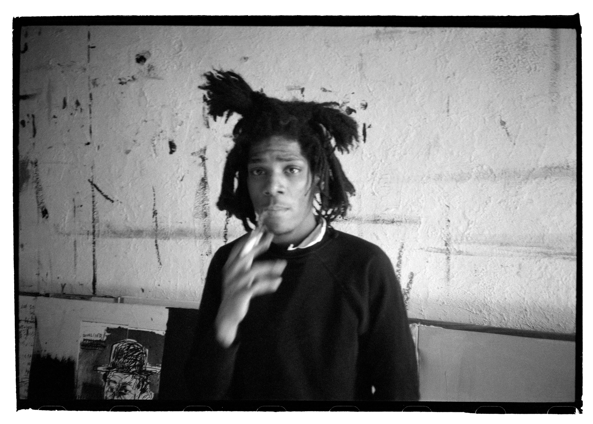 Fotografía en blanco y negro de Jean-Michel Basquiat de pie frente a una pared manchada, mirando a la cámara con las cejas levantadas y sosteniendo un cigarrillo en la mano.