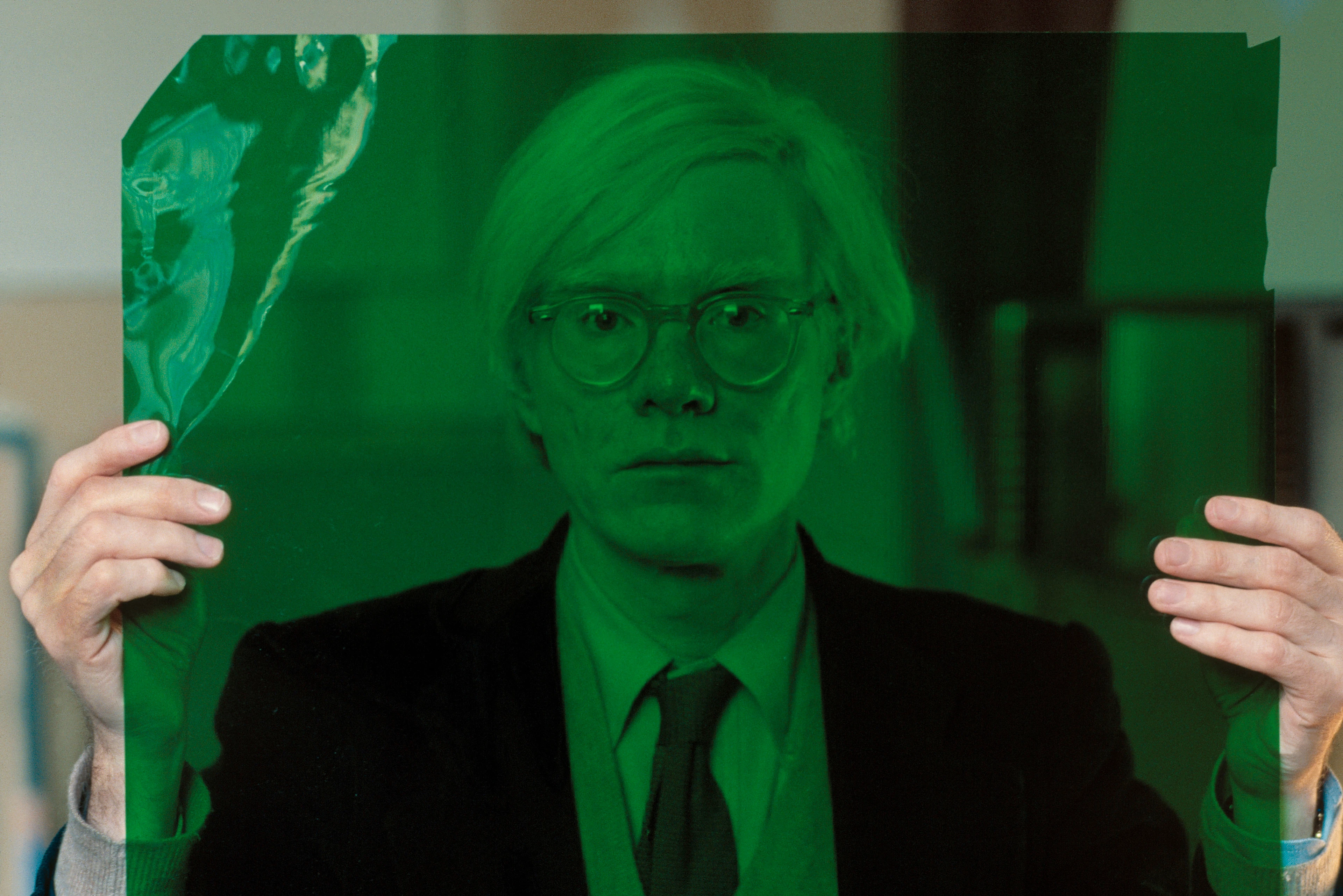 Retrato de Andy Warhol con traje y gafas, en el que mira a cámara y sostiene con ambas manos un papel de celofán verde delante de su rostro.