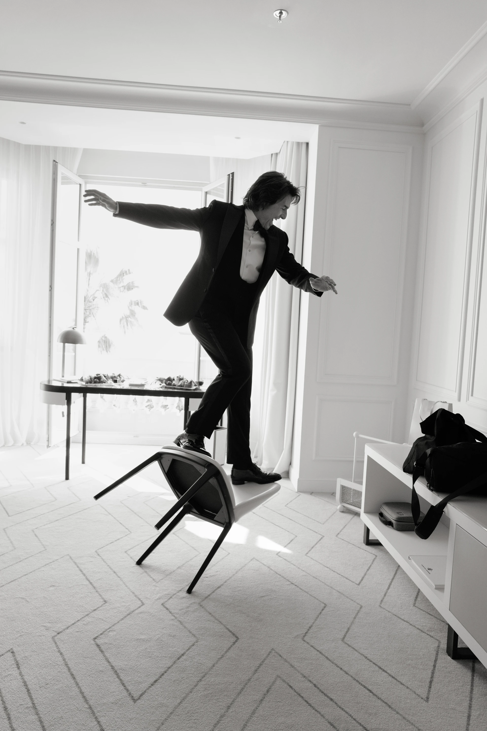 A man wearing a tuxedo playfully balances on a tilting chair in a bright hotel room with an open balcony door.