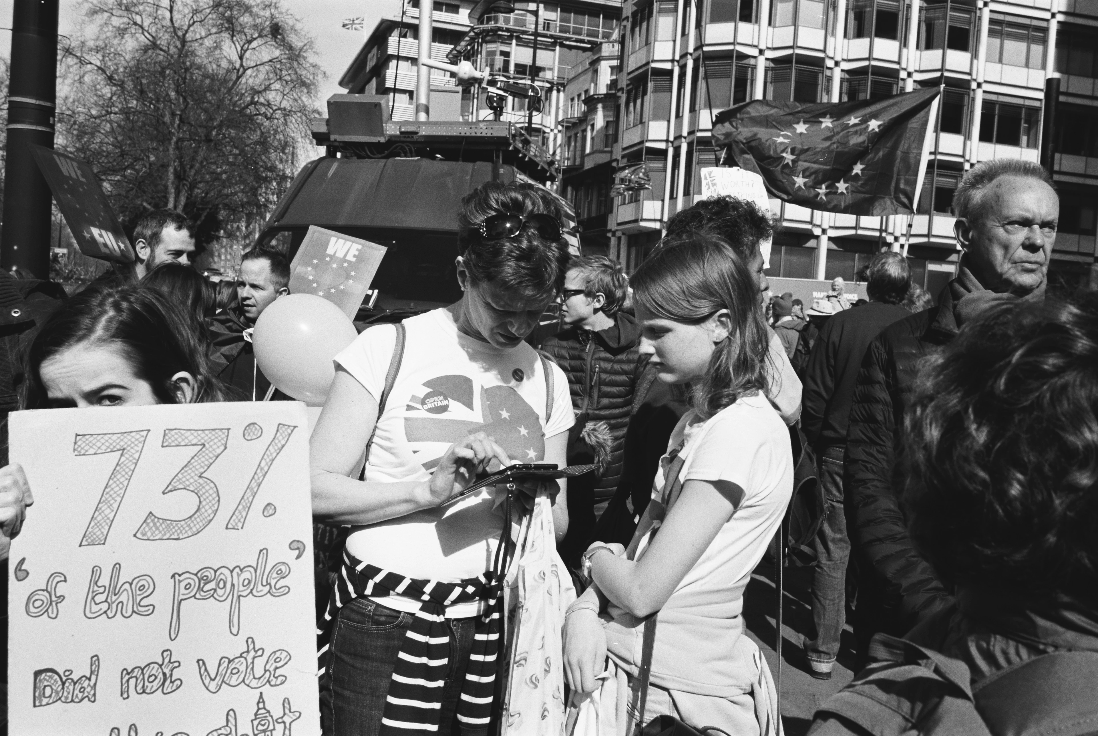 Black-and-white image: A woman surrounded by protesters types something into her smartphone while a younger woman looks on. A poster bearing the words ‘73% did not vote’ can be seen at the edge of the image.